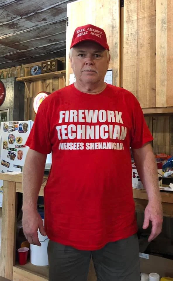 A man wearing a red T-shirt that reads 'Firework Technician Neesees Shenanigans' standing in a rustic wooden room.