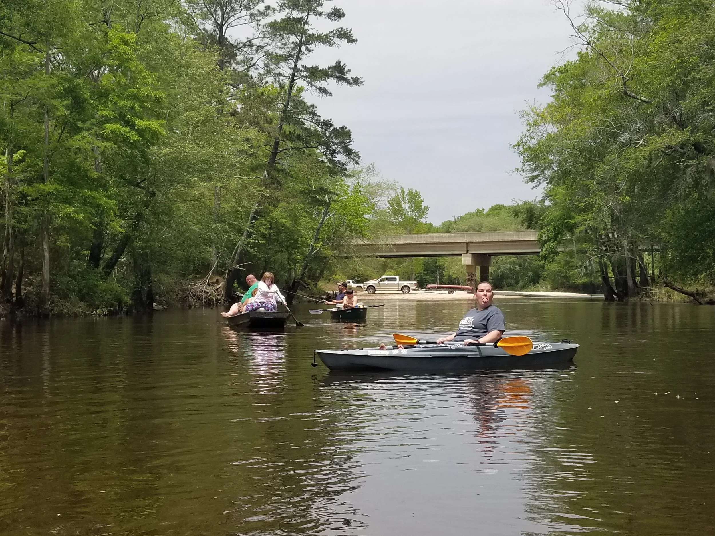 People kayaking and floating on a river surrounded by trees with a bridge overhead.