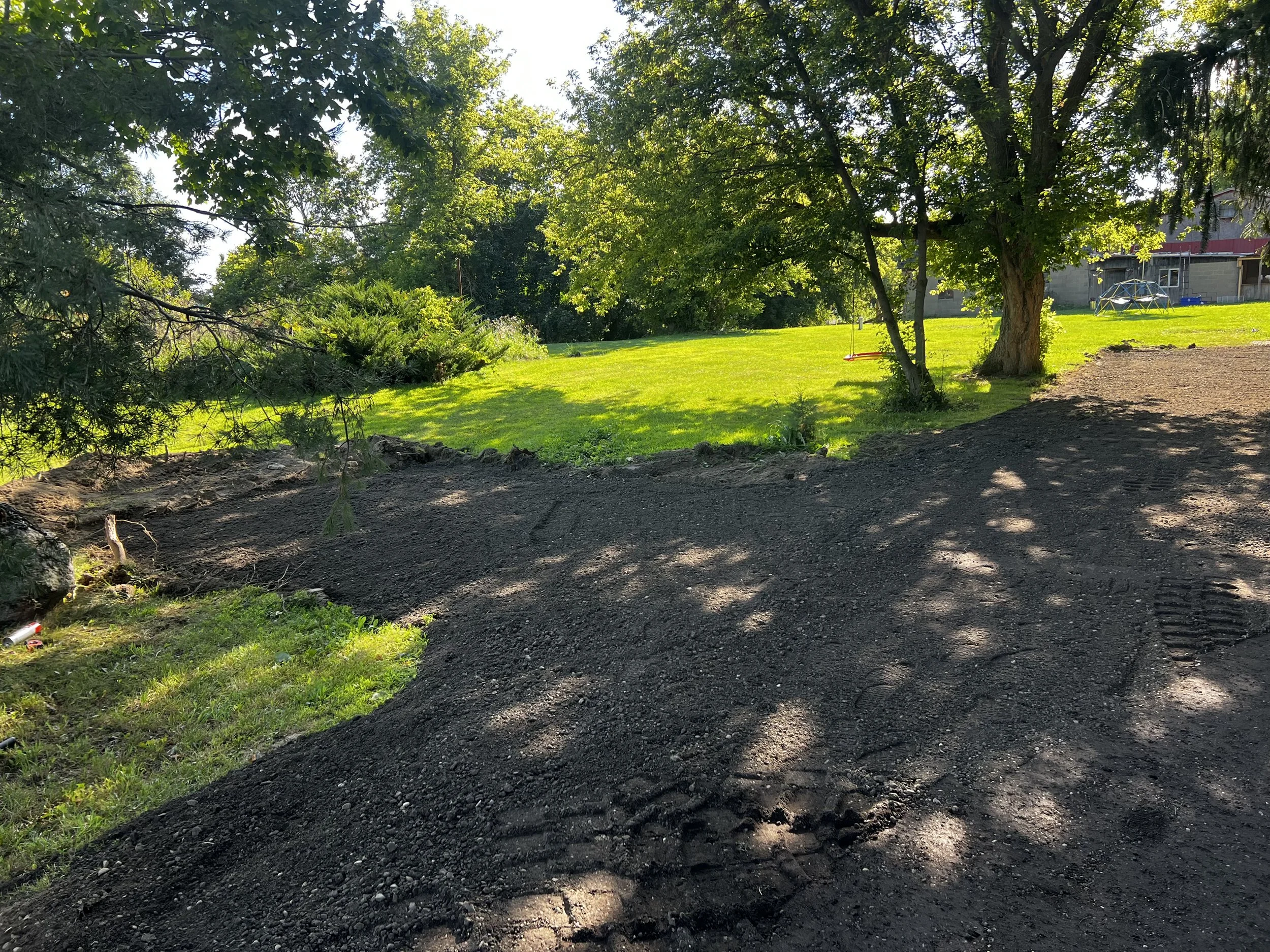 A backyard with a patch of freshly tilled dark soil, a large tree casting shadows, and a green lawn with some plants and a garden house in the background.
