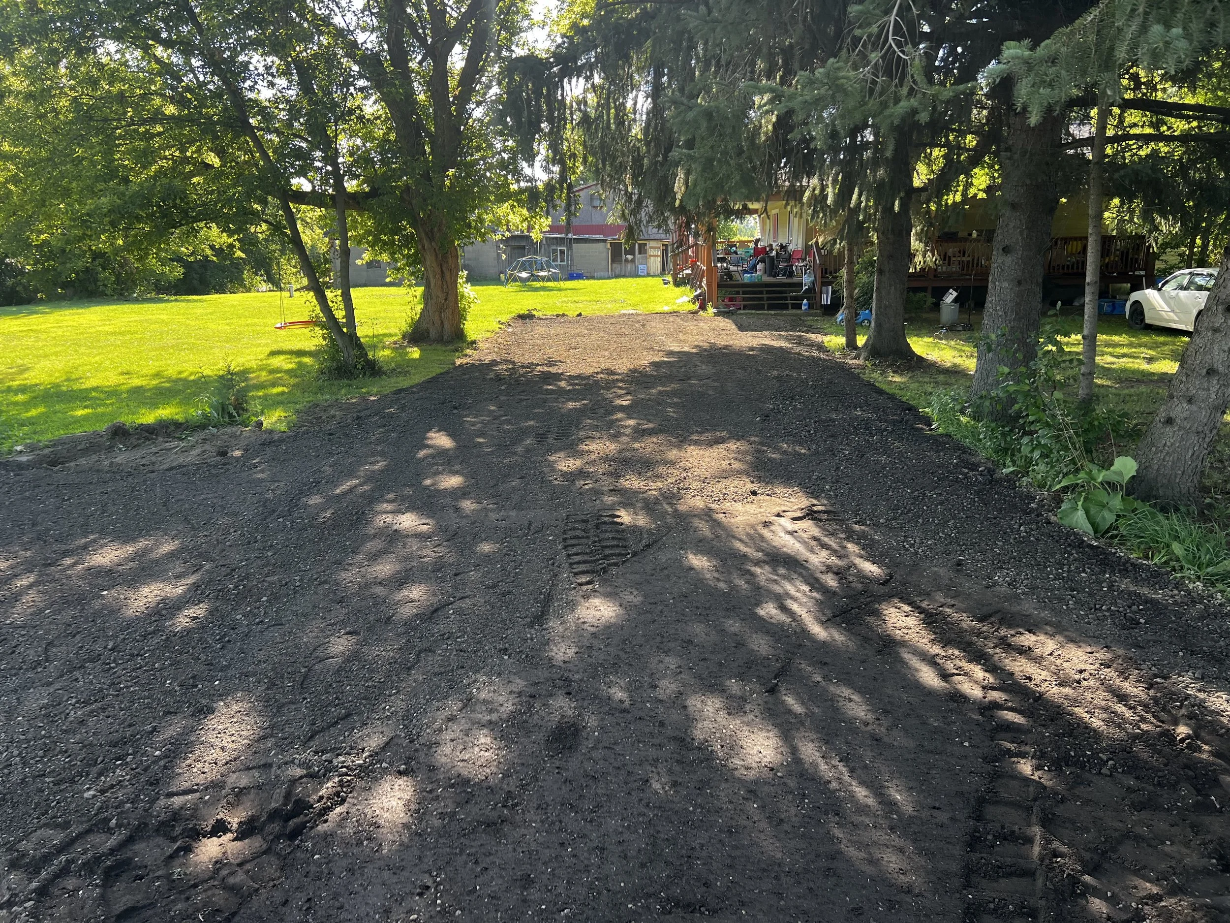 Une allée en gravier menant à une maison avec un porche en bois, entourée d'arbres avec des ombres projetées sur le sol.