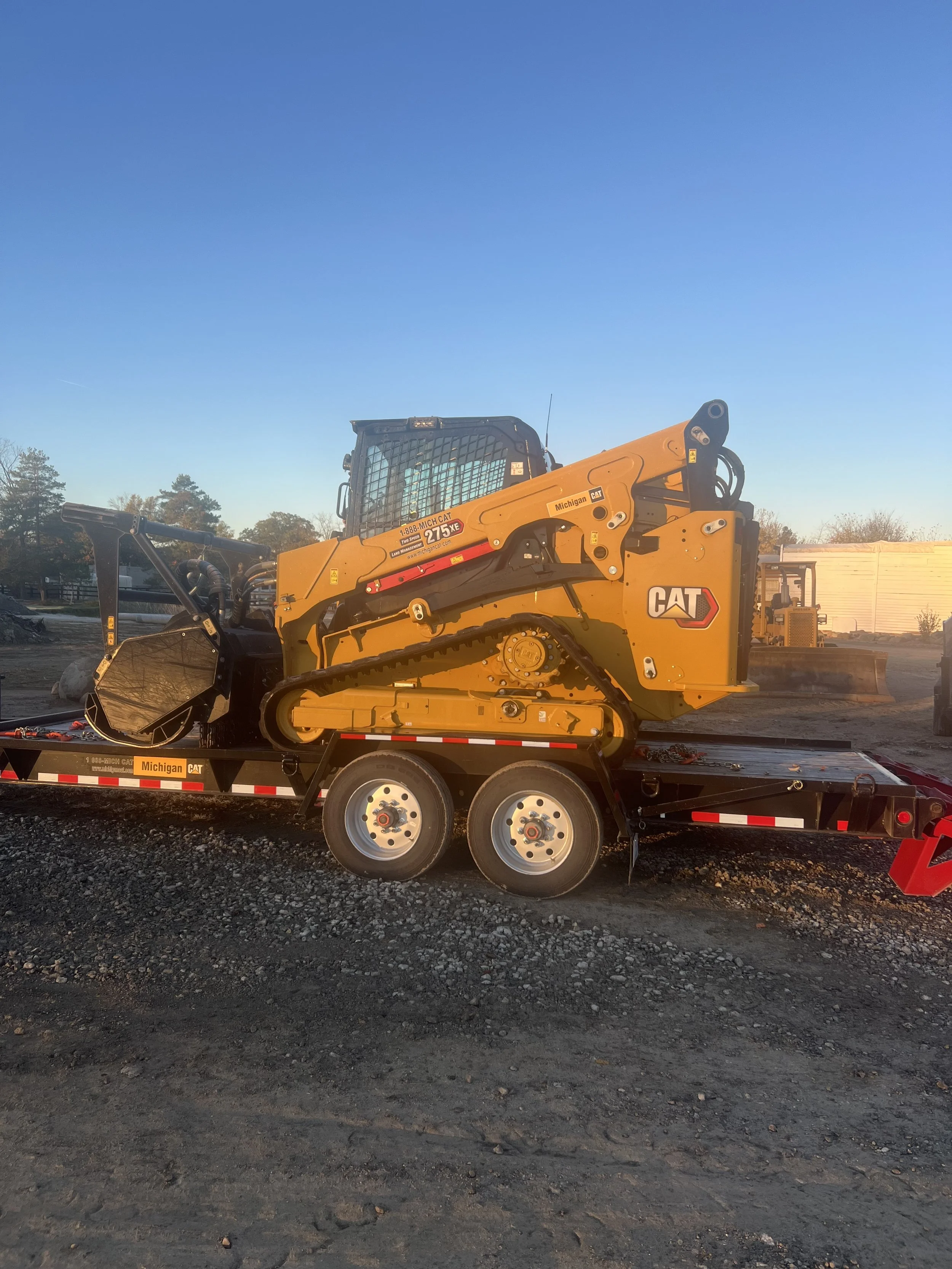 Yellow CAT skid steer loader loaded onto a black trailer in an outdoor lot with a clear blue sky and some trees in the background.