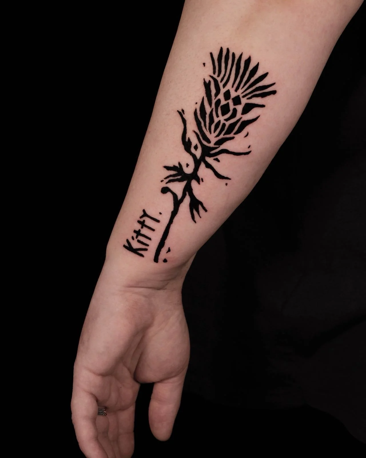 Bold lino-print floral tattoo on a person's forearm, with text vertically near the stem tattooed by Pip