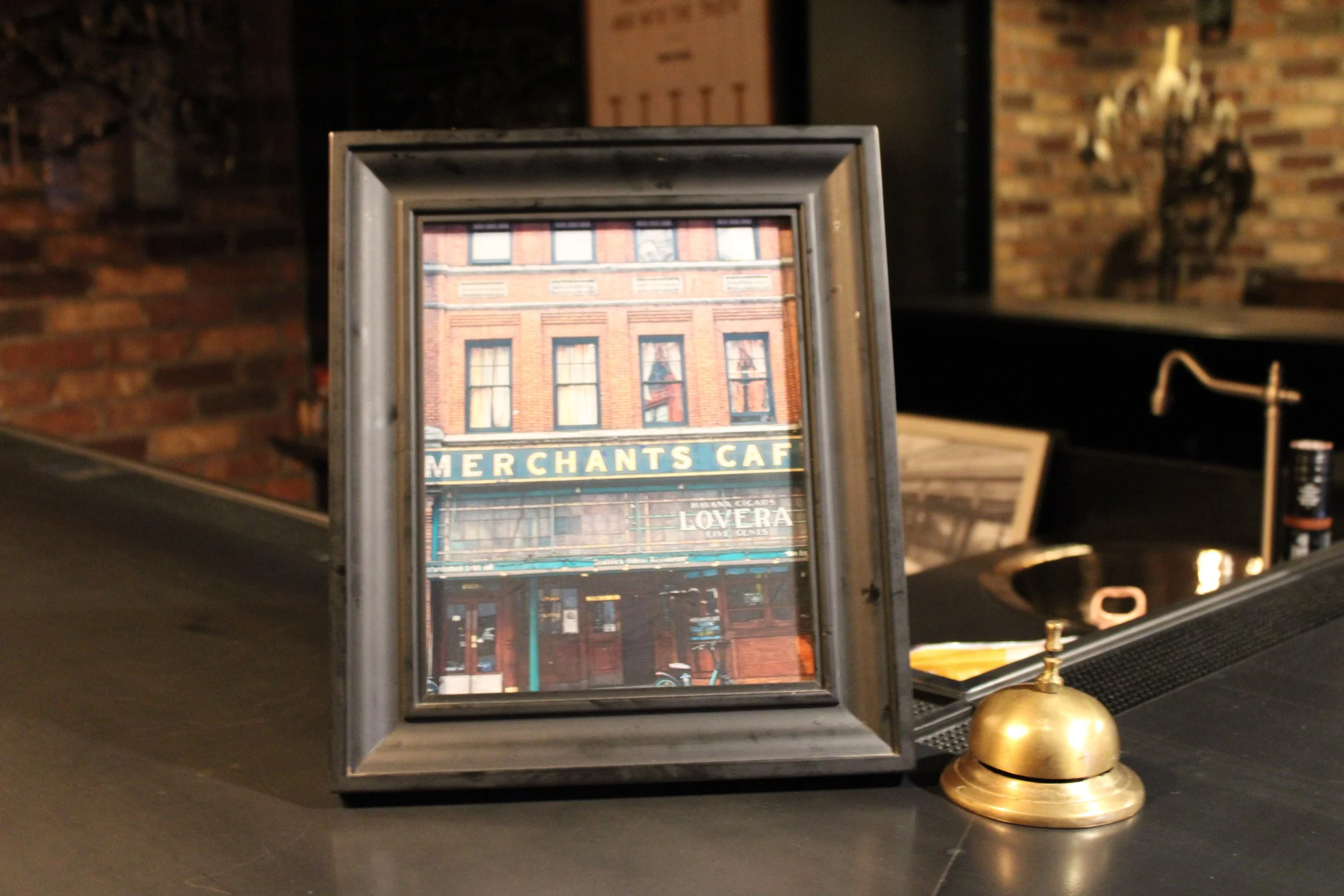 A framed photo of a building with a sign reading "MERCHANT'S CAFE" and a reflection of the same building in the glass, placed on a counter next to a brass object.