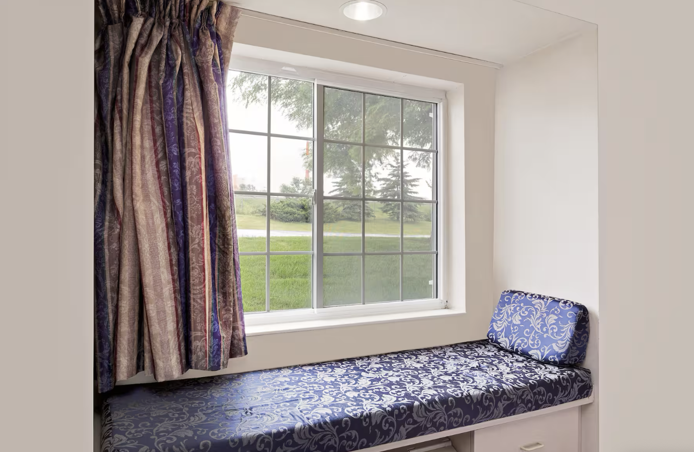 A window seat with a blue floral cushion and a matching footrest next to a large window with patterned curtains, overlooking a green lawn and trees outside.