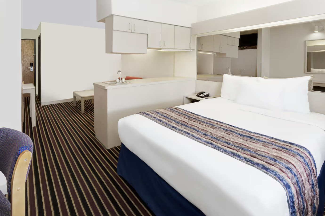Hotel room with a bed, striped carpet, kitchenette, and a small seating area.