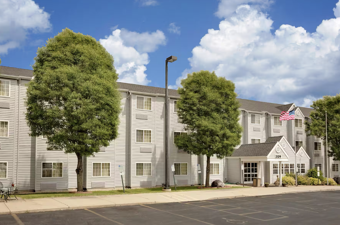 Exterior of a multi-story residential building with a grey siding facade, four levels, several windows, two large trees in front, an American flag, and a parking lot in the foreground.