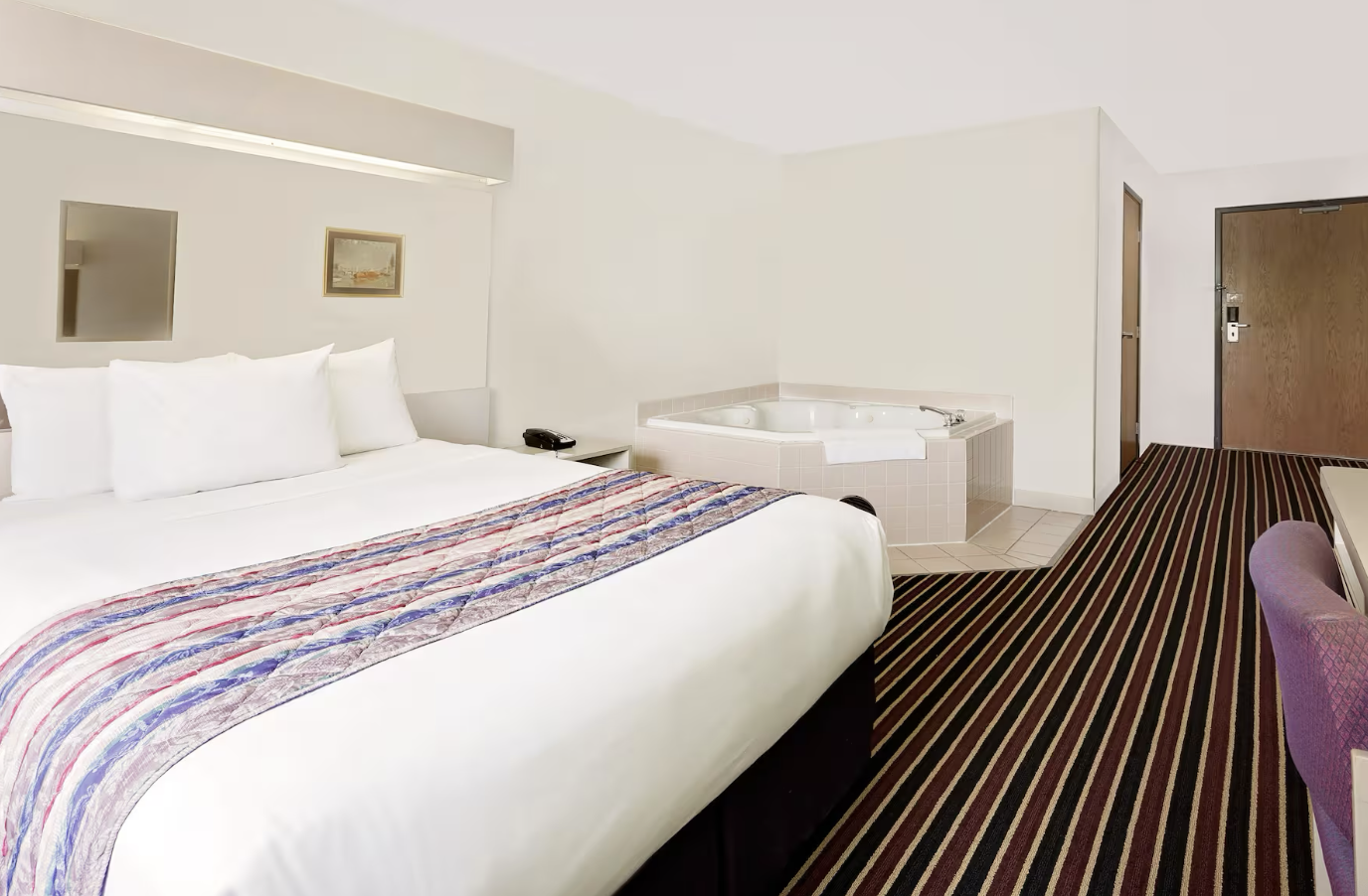 Hotel room with a large bed, bedside table, and bathroom jacuzzi in the corner. Carpeted floor and a wooden door.