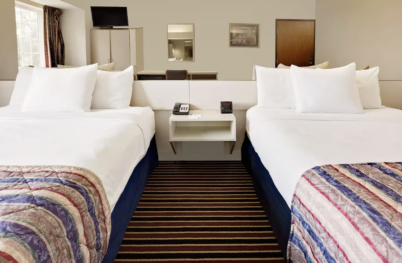 A hotel room with two beds, each covered with white bedding and multiple pillows, separated by a nightstand with a telephone and alarm clock. A striped carpet runs between the beds, and a window with curtains is visible on the left. A mirror, a small