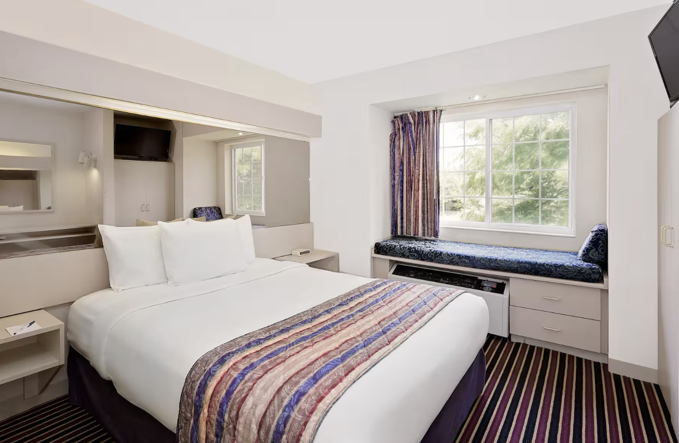 A hotel room with a large bed, window seat, striped carpet, and a window with curtains showing trees outside.