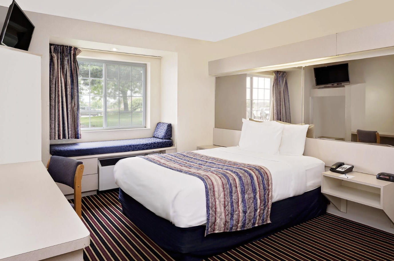 Hotel room with a bed, window seat, window with curtains, TV, and desk, decorated in neutral tones with striped carpet.