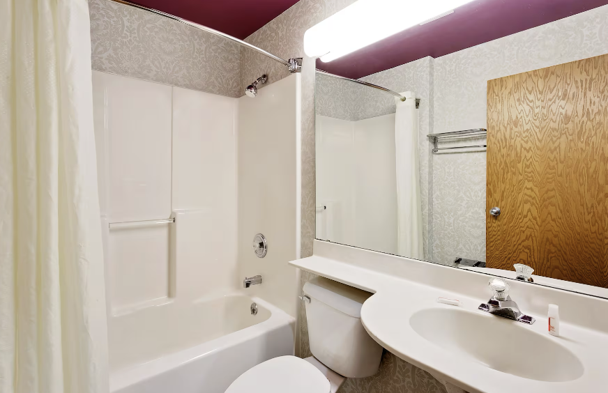 Small bathroom with a bathtub, shower curtain, toilet, and a sink with a large mirror above. There is a wood door and a towel rack on the wall. The ceiling has a fluorescent light fixture.