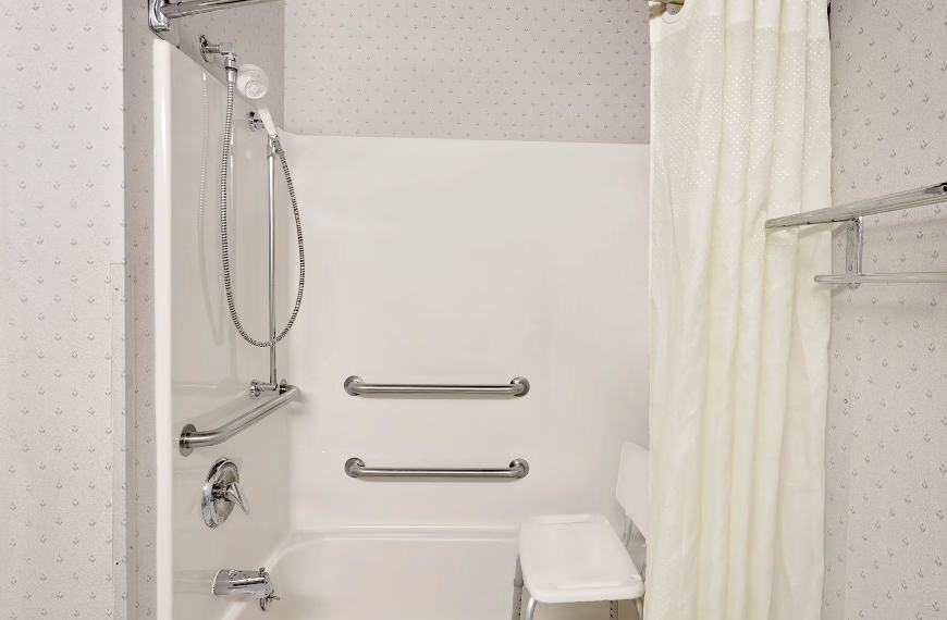 Accessible shower with grab bars, a handheld shower head, and a folding chair, partially covered by a shower curtain in a small bathroom.