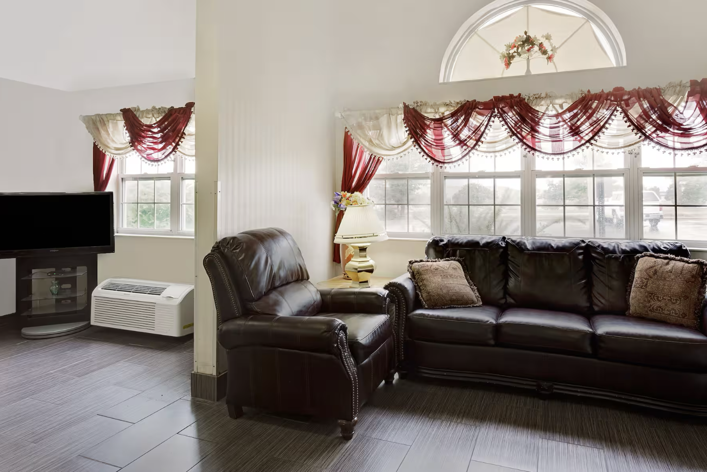 Living room with large windows decorated with red and white curtains, a dark brown leather armchair and sofa, a table lamp with a floral arrangement, a TV, and an air conditioner.