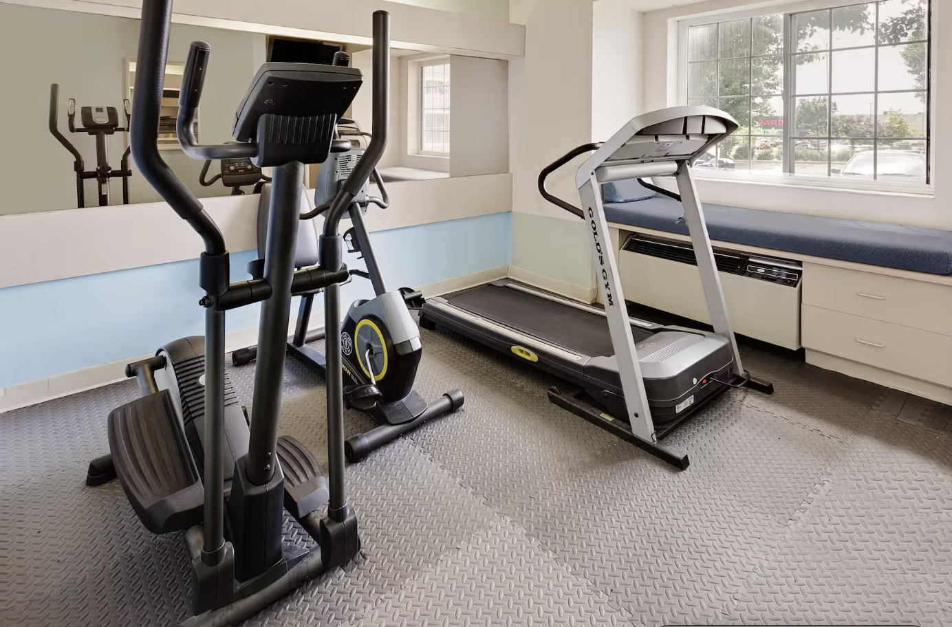 Room with exercise equipment including an elliptical machine and a treadmill, beside a large window with street view.