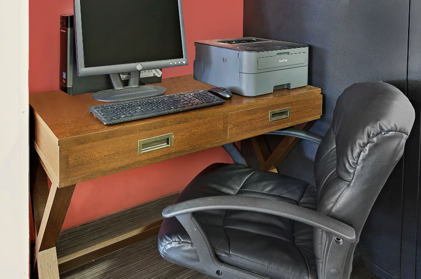 A workspace with a wooden desk, a computer monitor, keyboard, mouse, and a printer, alongside a black leather office chair.