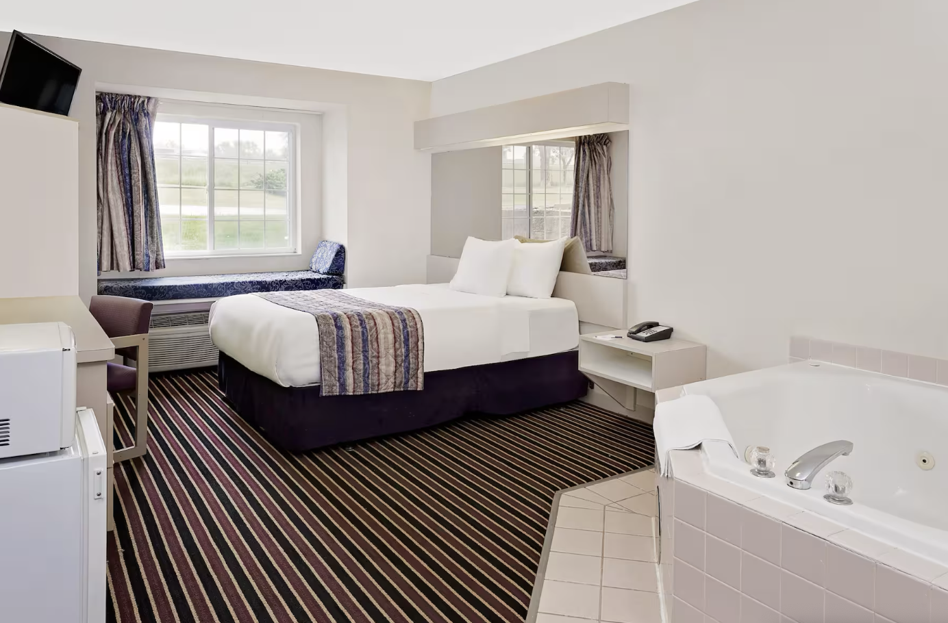 A hotel room with a bed, window with curtains, a window seat, a chair, a small refrigerator, a microwave, a wall-mounted TV, a telephone, and a bathtub.