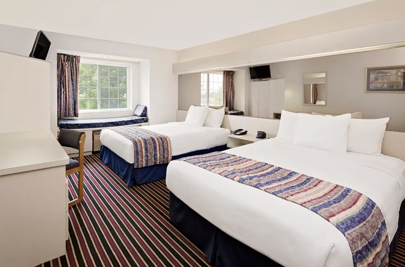 Hotel room with two beds, window seat, striped carpet, and wall-mounted TVs.