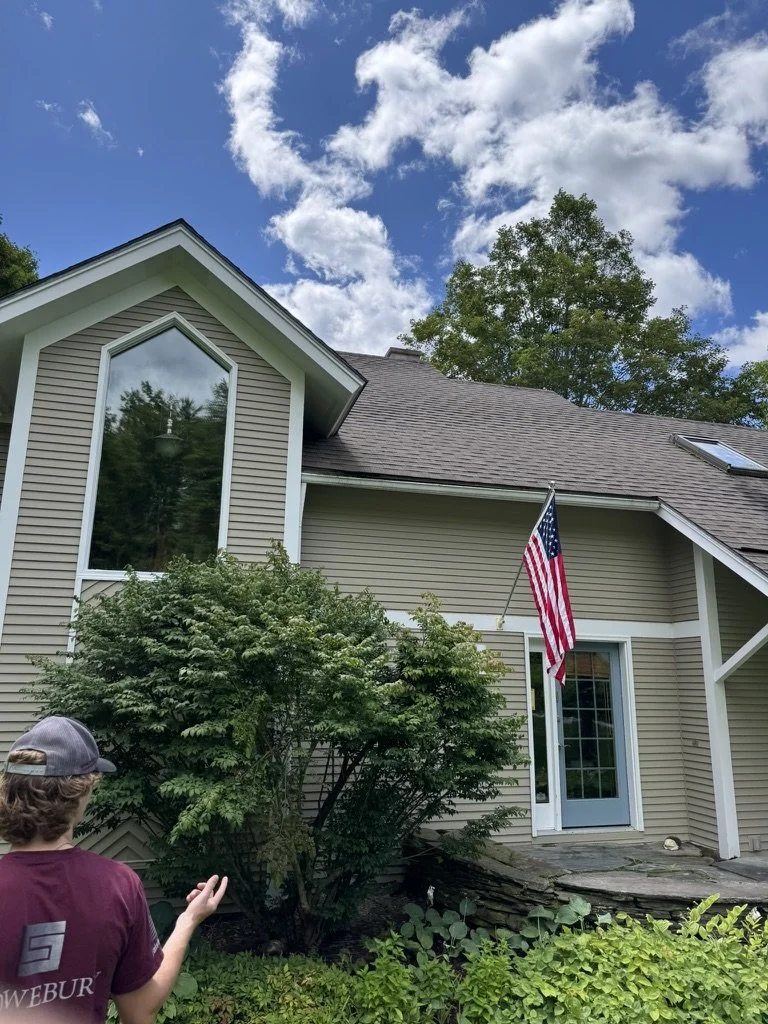 A house with beige siding and a triangular window on the upper floor, an American flag hanging near the door, lush green bushes in front, a person in a maroon shirt and cap standing nearby, and a partly cloudy blue sky above.