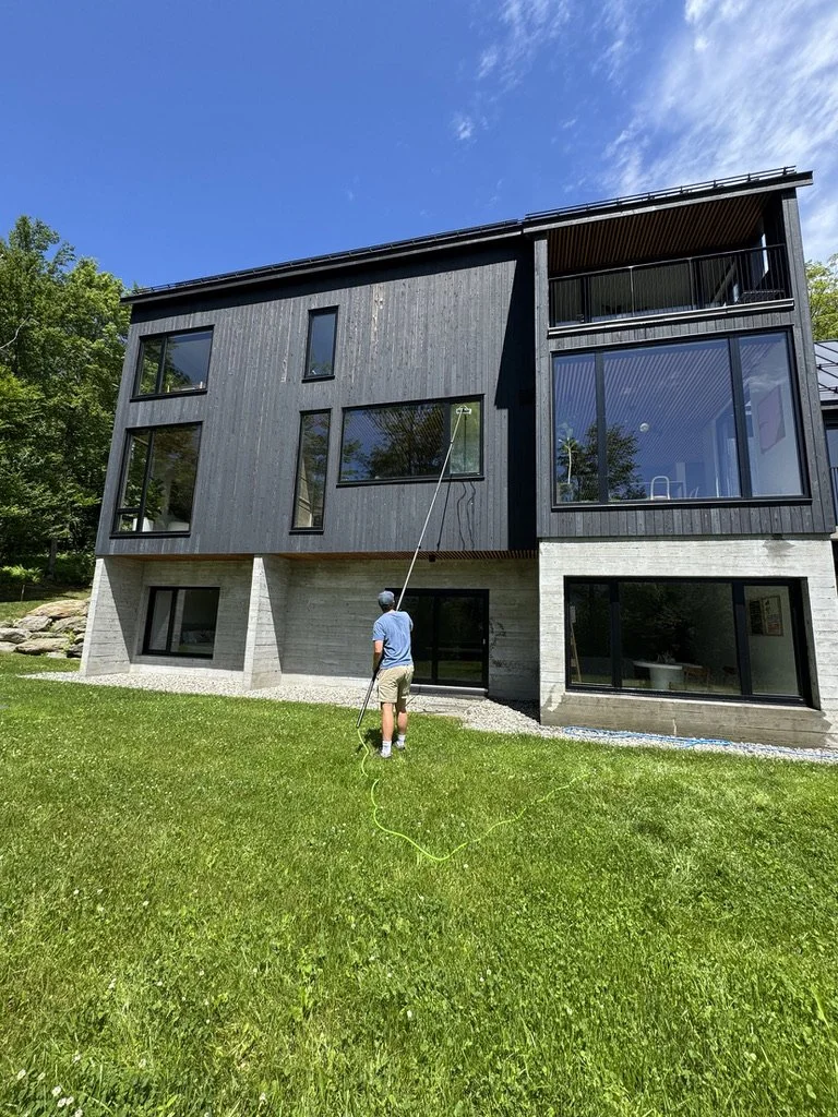 A person is using a long pole to spray or clean the exterior windows of a modern house with black siding and large glass windows, located on a grassy yard with trees in the background under a blue sky.