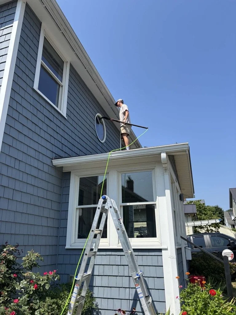 A man cleaning a round window on the second floor of a blue house using a long-handled tool, standing on the roof attached to a ladder, under a clear blue sky, with plants and flowers below.