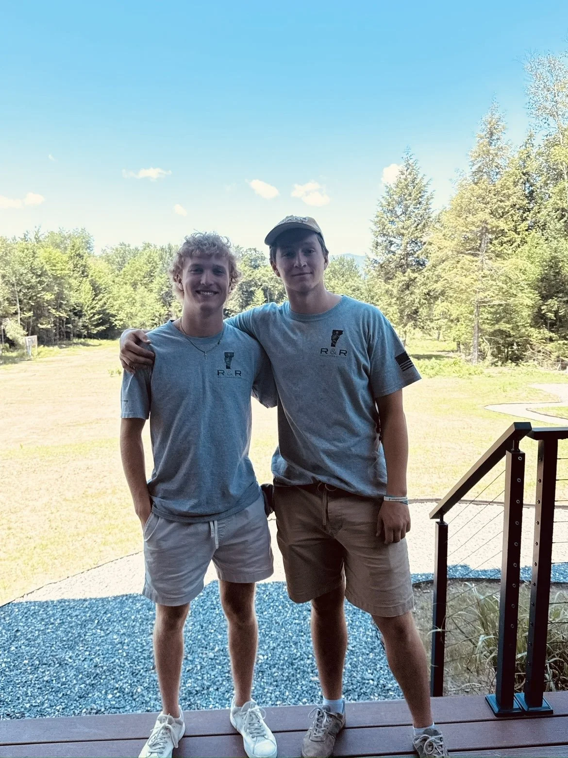 Two young men standing outdoors, smiling, with trees and grass in the background. They are wearing matching light grey T-shirts and khaki shorts.