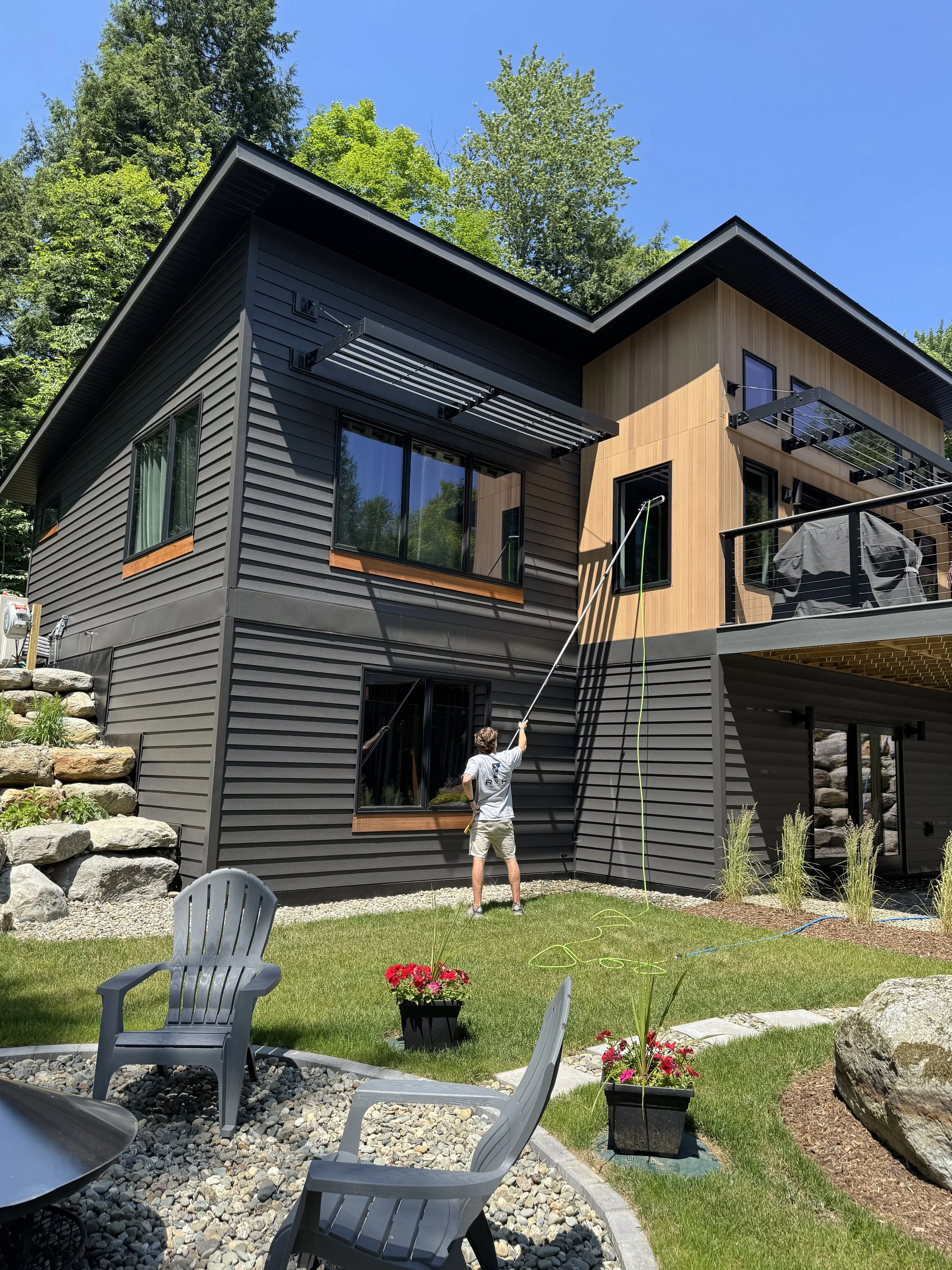 A man standing outside on a backyard patio, spraying water onto the house with a hose. The house has black and natural wood siding, large windows, and small patio awnings. There are landscaped plants, a stone border, and patio furniture with potted flowers under a clear blue sky.