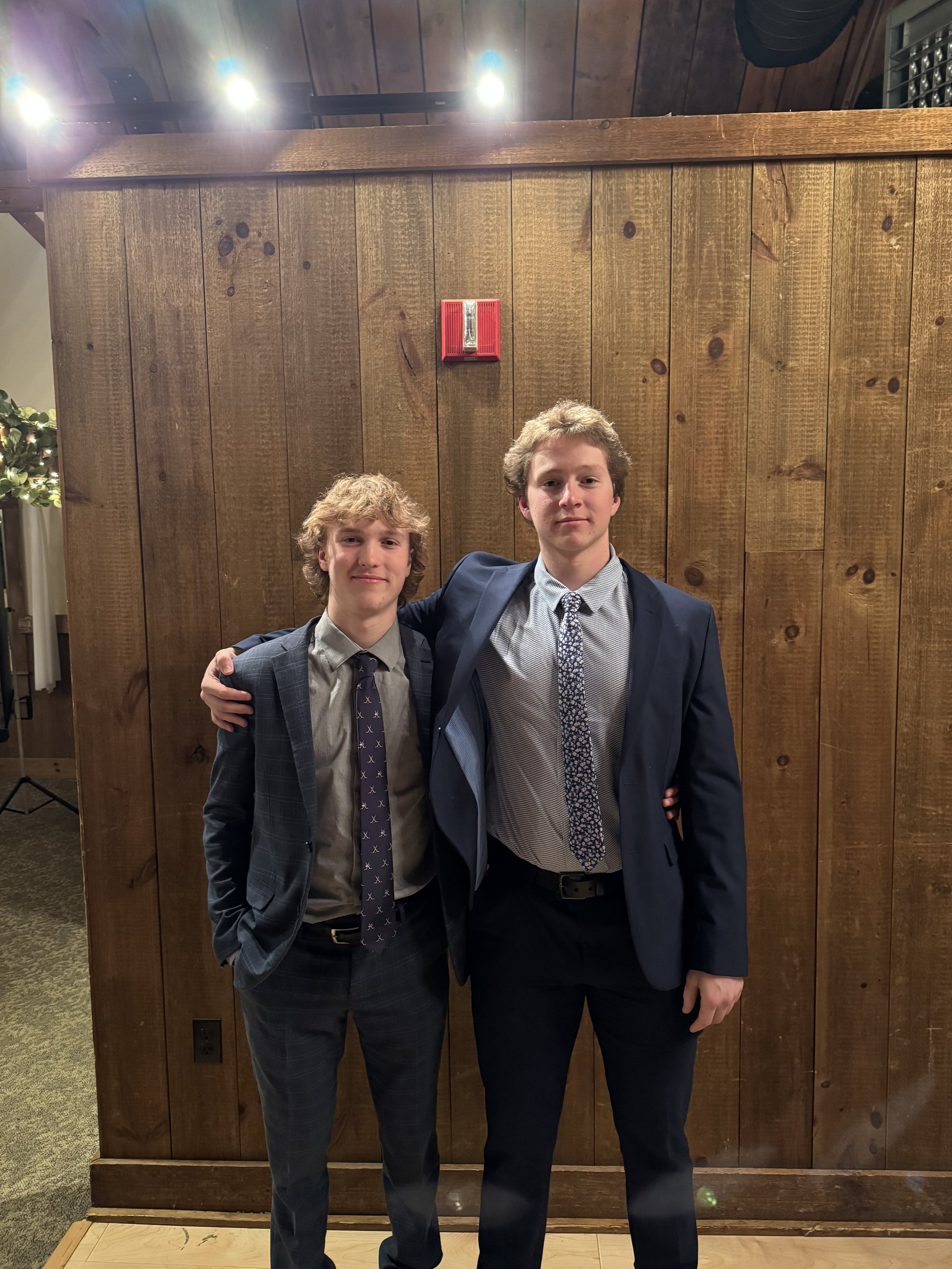 Two young men in suits standing together in front of a wood-paneled wall, with one arm around each other's shoulders, smiling at the camera.