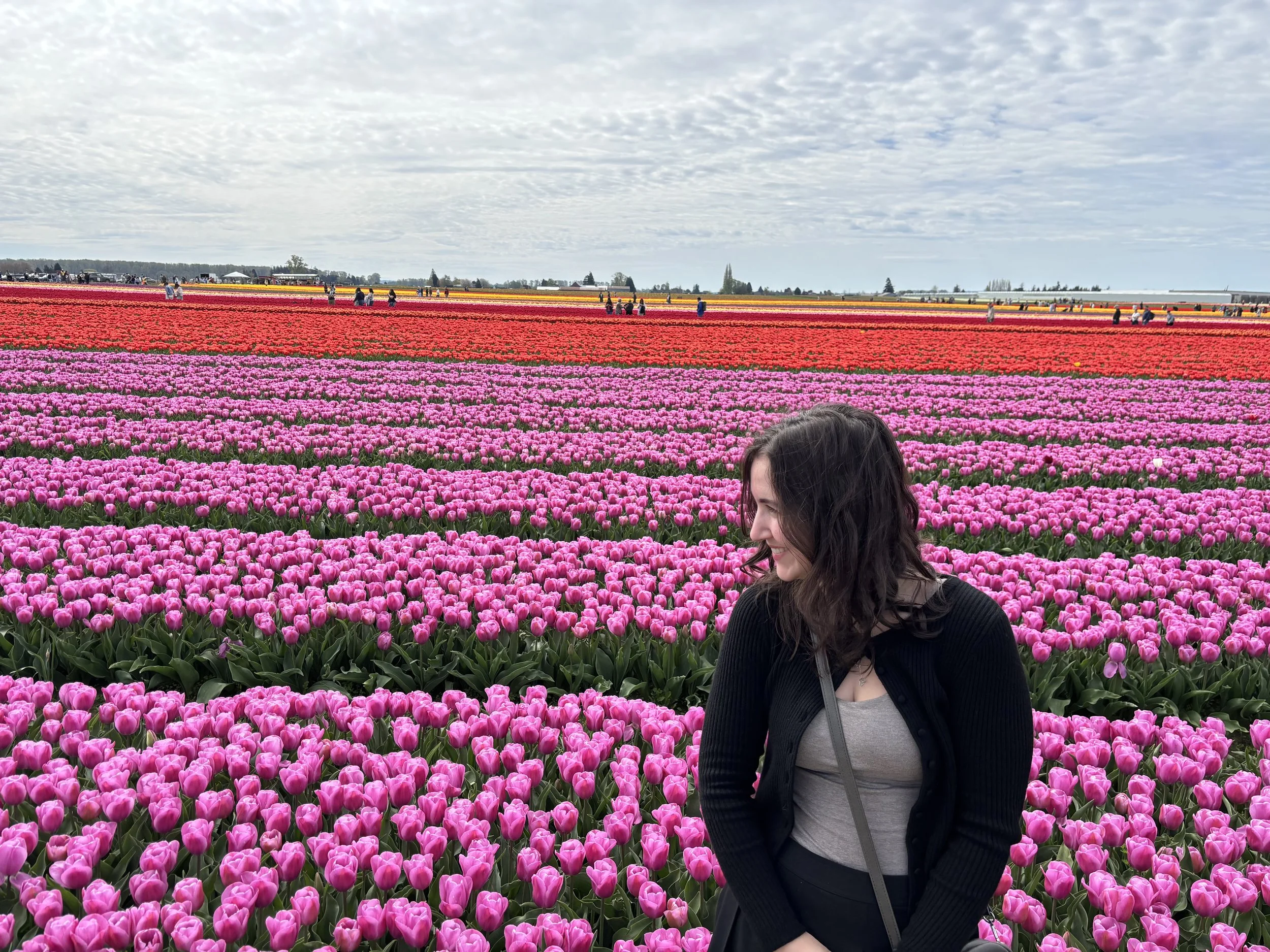 A woman with dark hair in a black jacket and gray shirt standing in a vast field of pink, red, and yellow tulips under a cloudy sky.