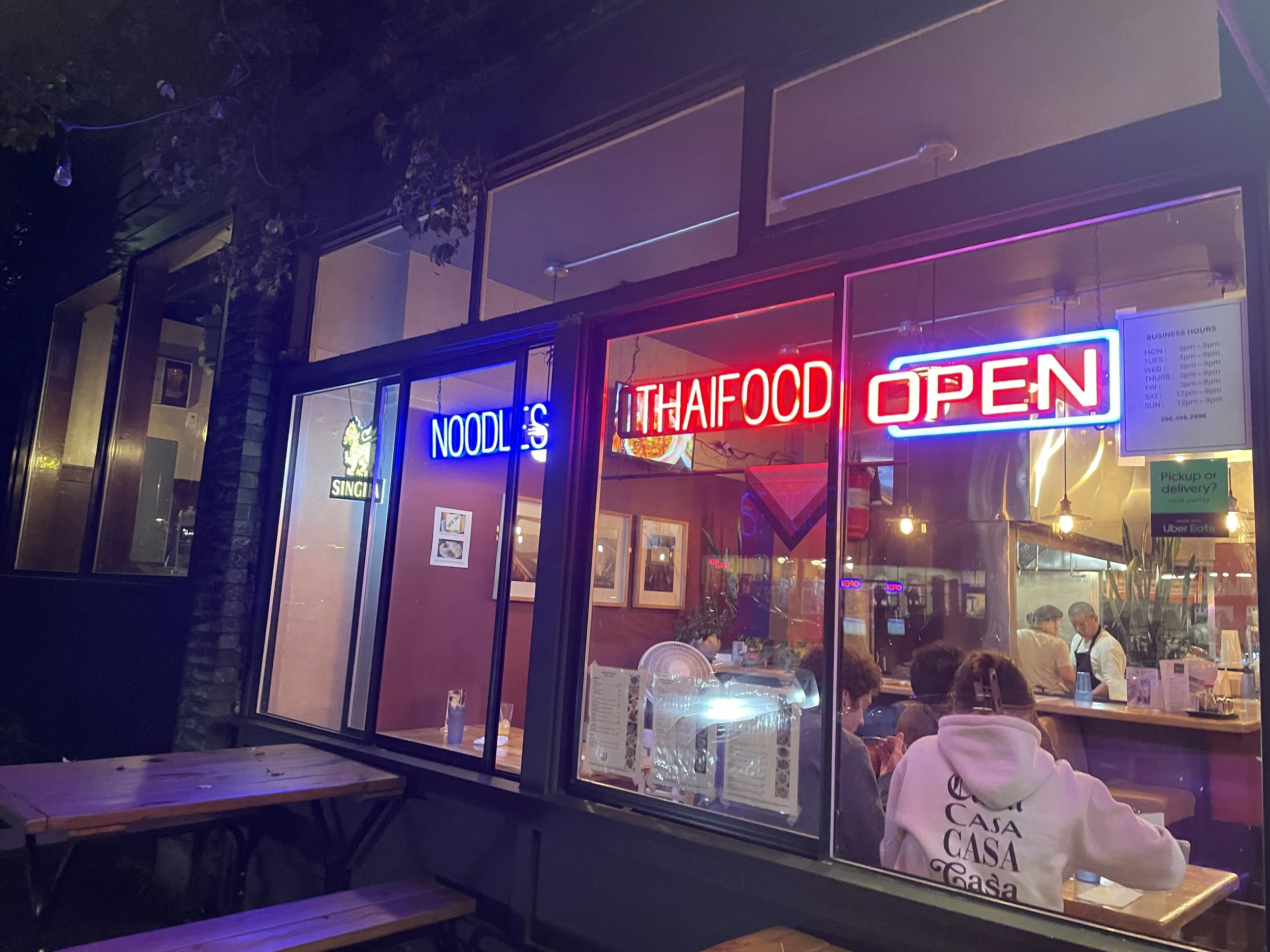 Nighttime view of a restaurant with neon signs reading 'THAI FOOD' and 'OPEN', inside with customers seated, and a window reflecting lights and trees outside.