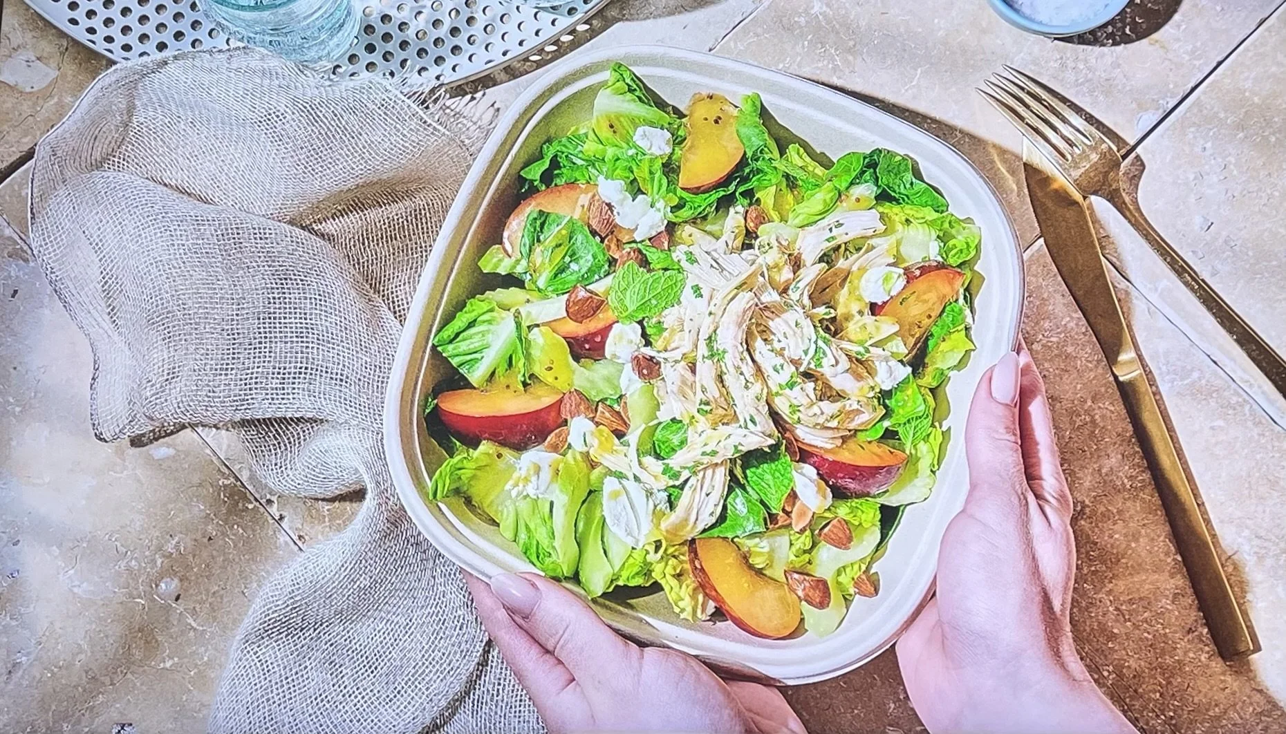 A person holding a bowl of fresh salad with mixed greens, sliced peaches, nuts, and cheese, on a dining table.