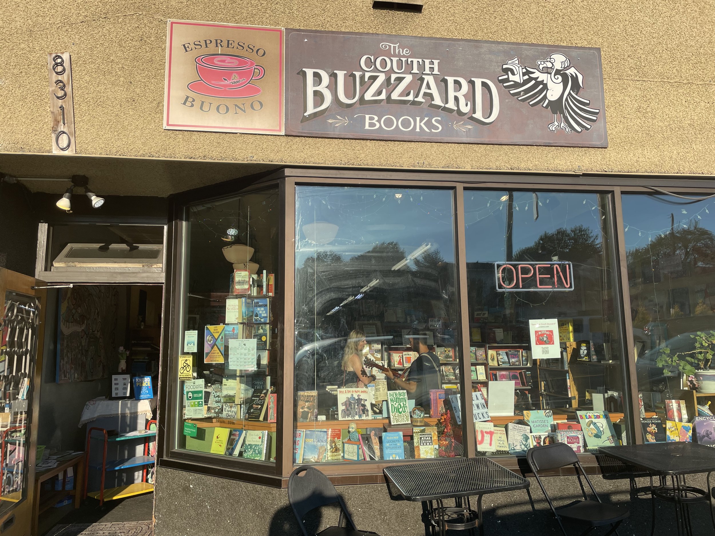 Bookstore storefront with signs reading 'The South Buzzard Books' and 'Espresso Buono.' Large window displays books, and there are tables and chairs outside under a sunny sky.