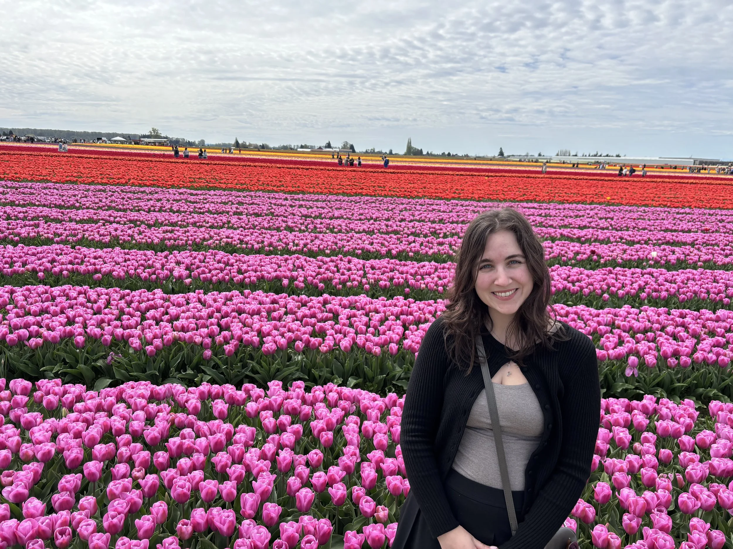 A woman with brown hair and a black jacket smiling at the camera in a colorful tulip field with pink, red, orange, and yellow tulips and cloudy sky.