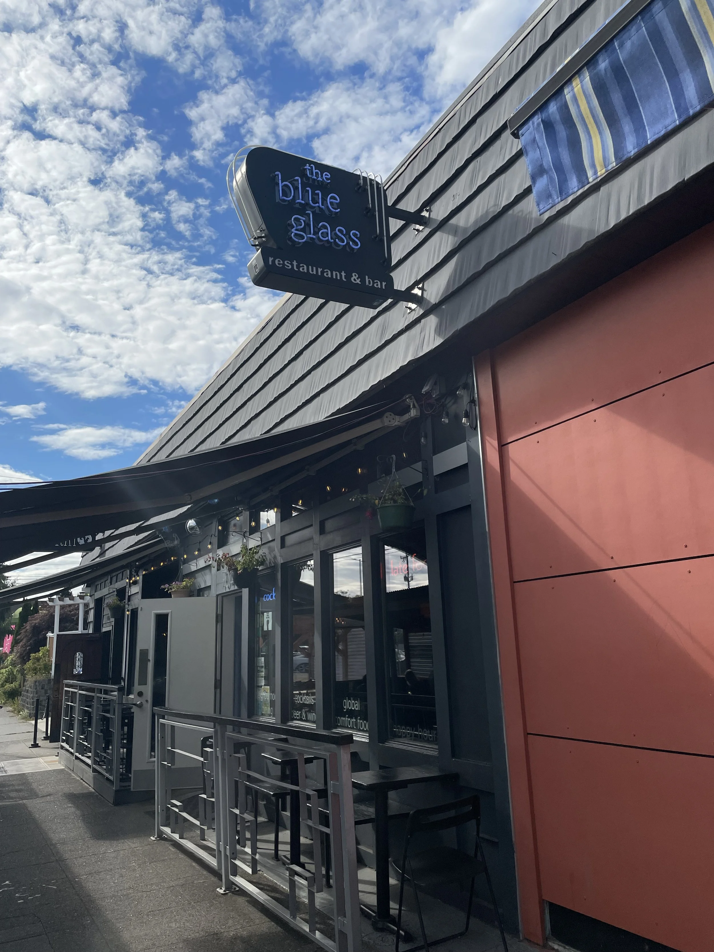 Exterior view of a restaurant and bar called 'the blue glass' with outdoor seating and a blue and yellow striped awning, under a partly cloudy sky.