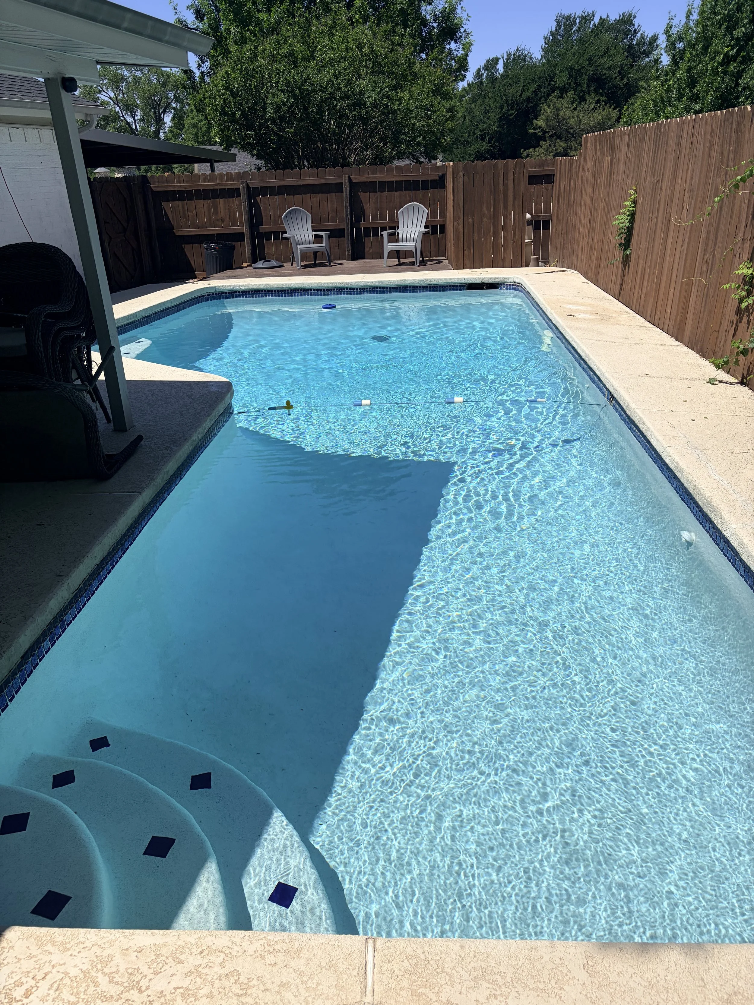 A backyard swimming pool with clear blue water, surrounded by a concrete deck, with two white plastic chairs on the back deck and a wooden privacy fence.
