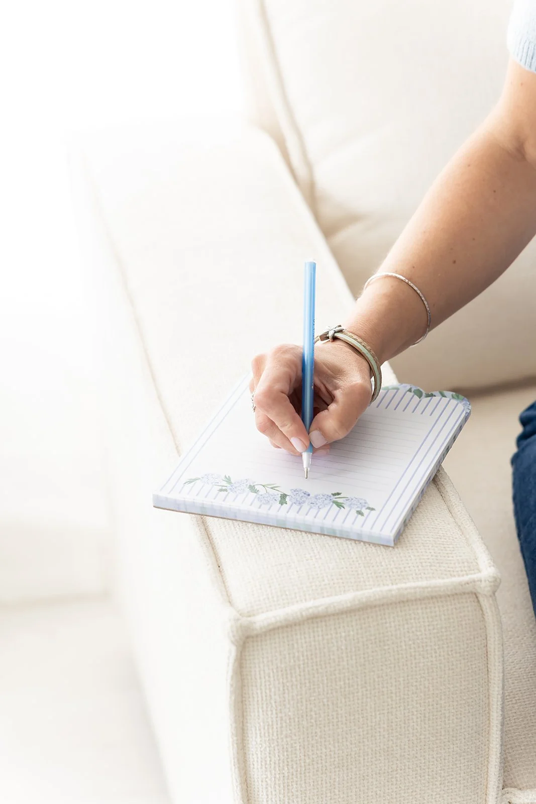 Person writing in a small floral notepad on a cream-colored sofa with a light background.