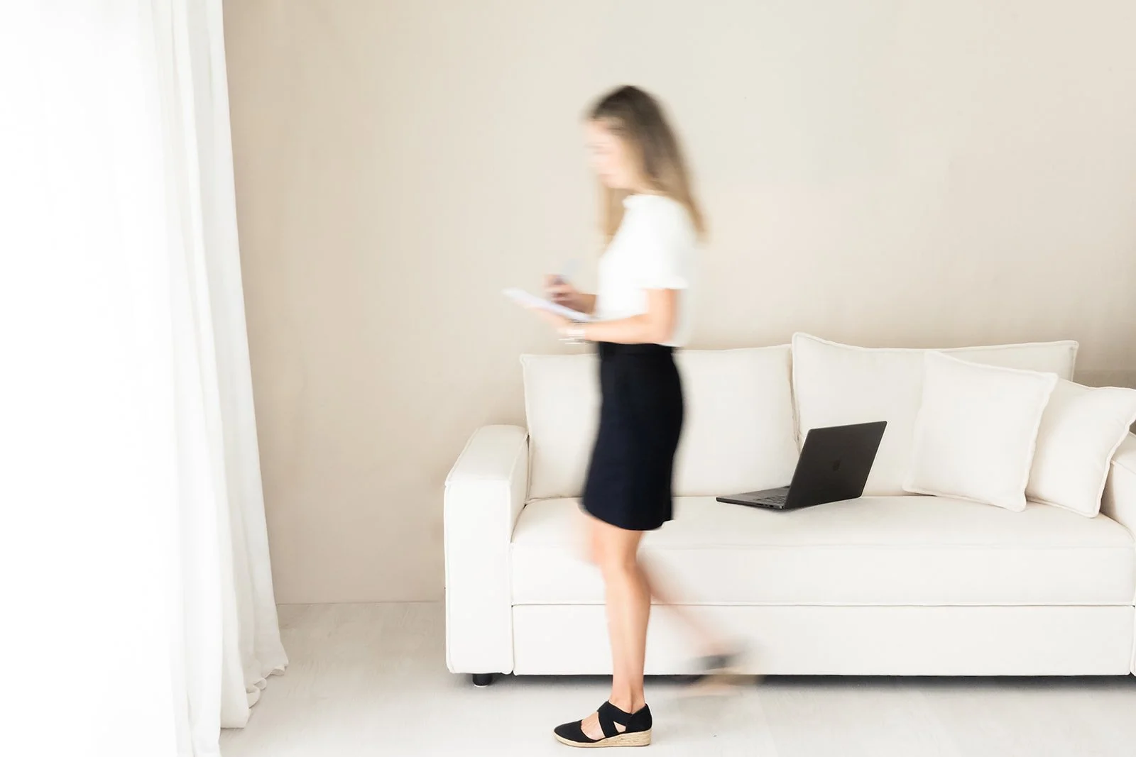 A therapist walking past a white sofa with a black laptop on it, holding a phone, in a bright minimalist room with white walls and curtains.