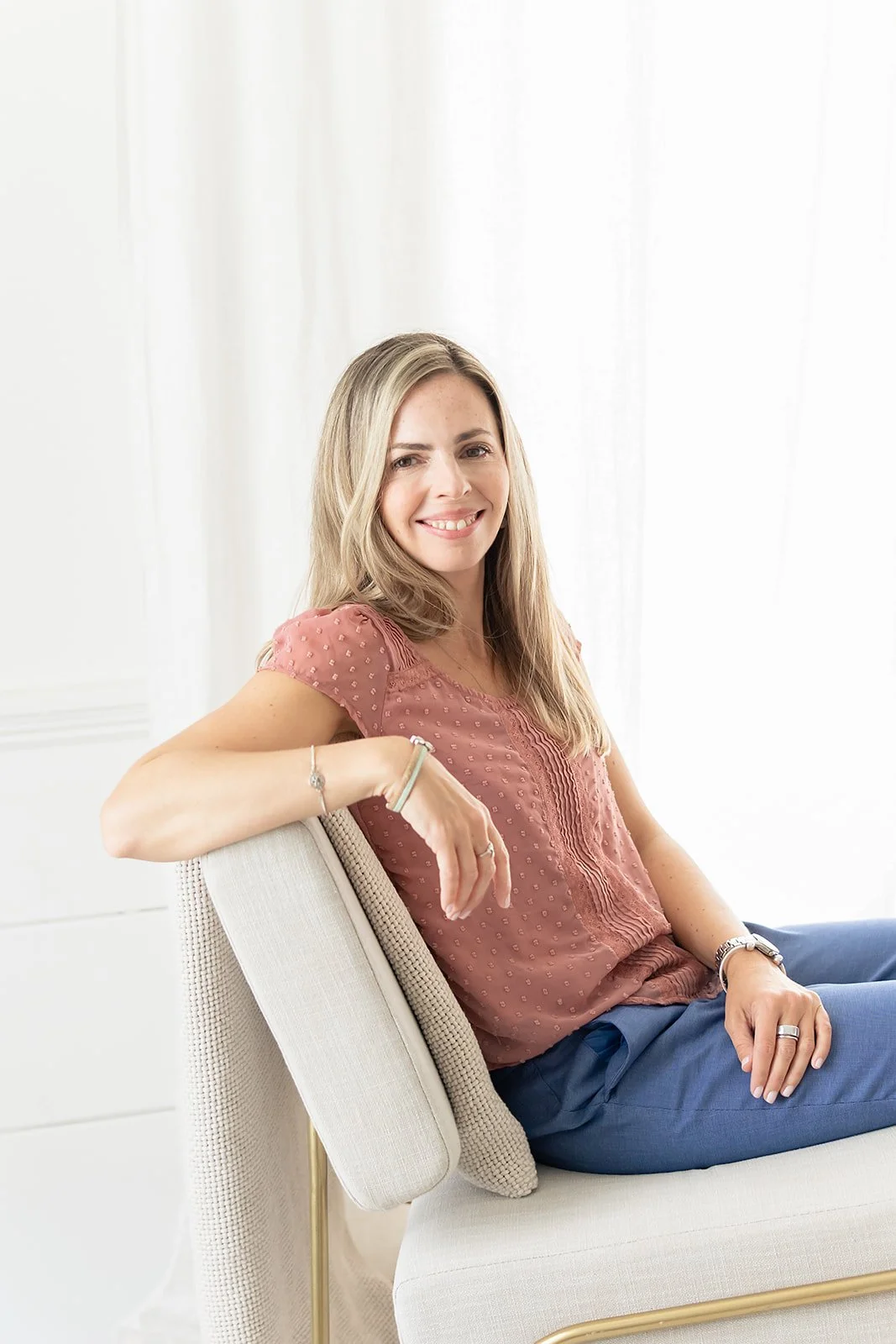 A therapist with long blonde hair wearing a pink blouse and blue pants, sitting on a beige chair indoors with white curtains in the background.