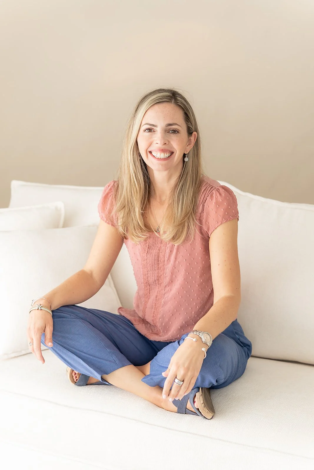 A therapist with long blonde hair, wearing a pink blouse and blue pants, sitting cross-legged on a white sofa, smiling.