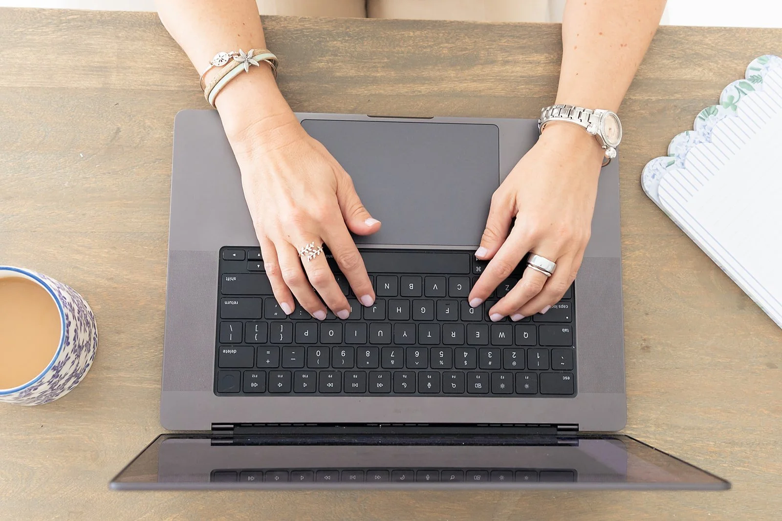 Top-down view of a woman typing on a laptop keyboard at a wooden desk. She is wearing a silver watch and several rings, with a coffee mug on her left and a notebook on her right.