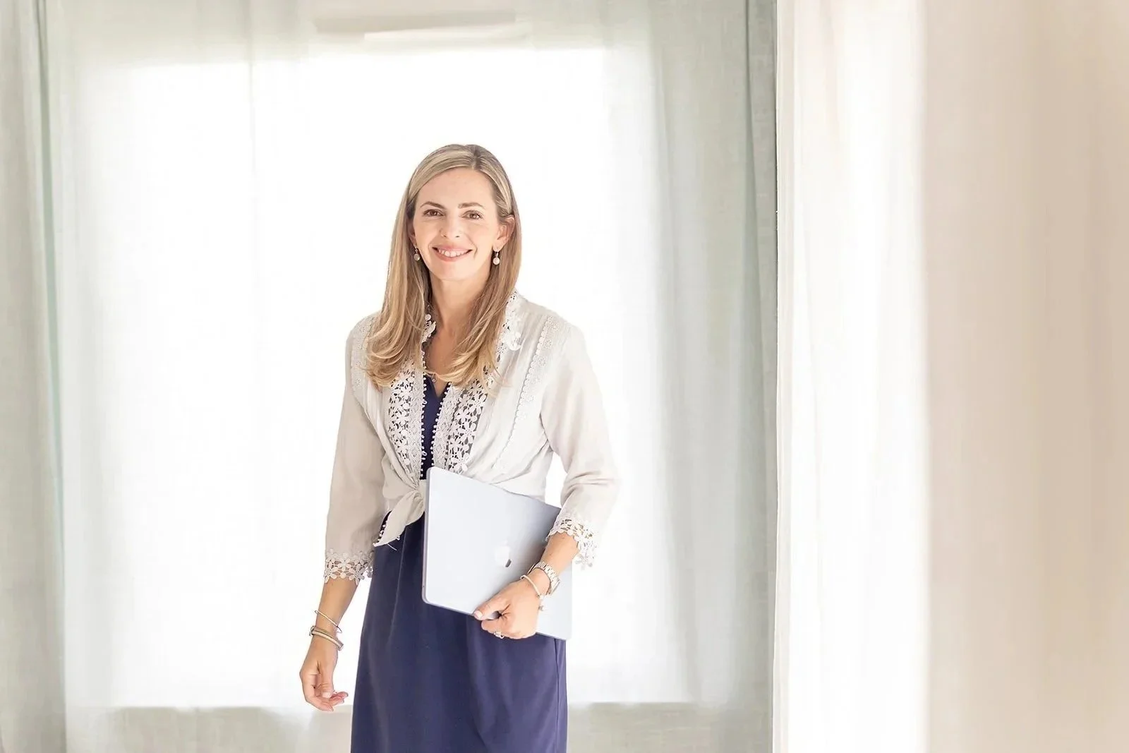 A Therapist with blonde hair and a smile, wearing a white lace blouse and a dark blue dress, holding a silver laptop and standing in front of a bright window with white curtains.