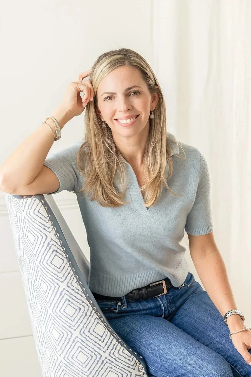 A woman with blonde hair smiling, sitting on a patterned chair, wearing a light blue sweater, blue jeans, and jewelry in a bright room.