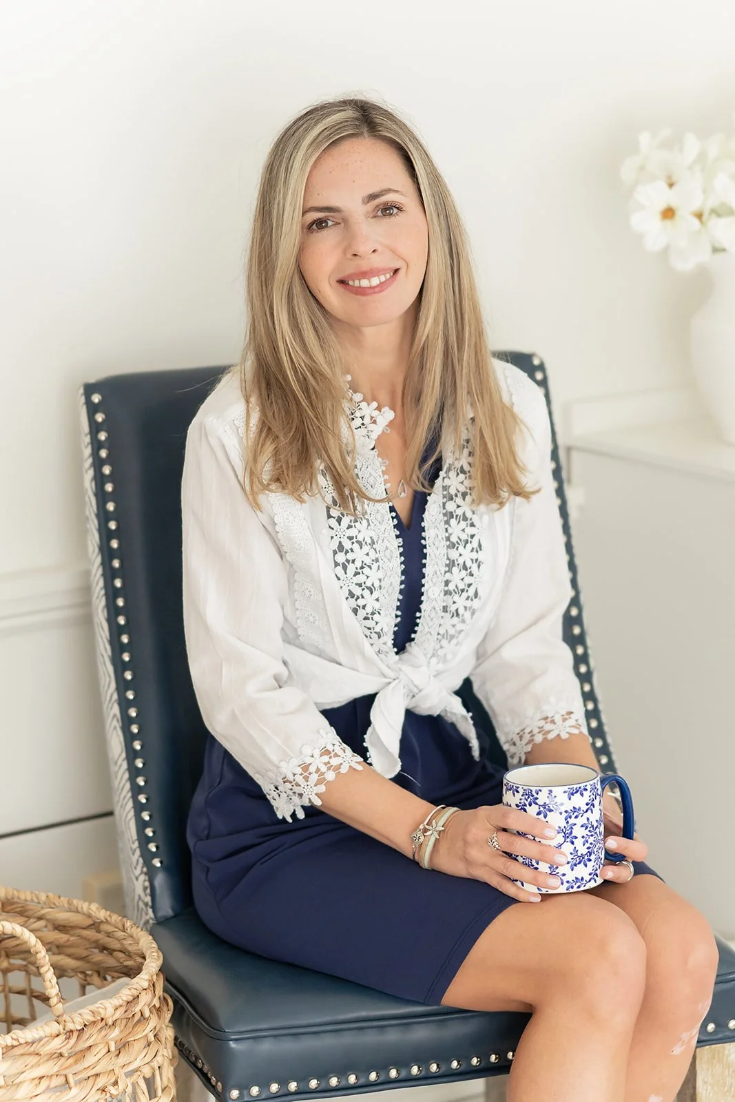 A therapist with blonde hair sitting on a navy blue chair in a room with white walls, holding a white mug with blue floral pattern, wearing a white embroidered blouse and navy skirt, with a vase of white flowers on a nearby table.
