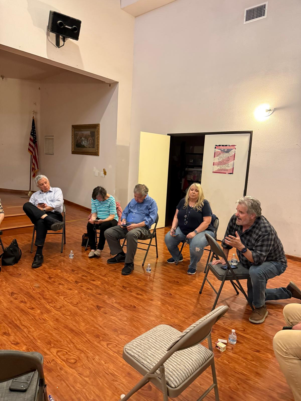 A group of five adults sitting on chairs in a circle in a room with wooden floors, engaged in conversation. Two women and three men, worshipping. An American flag is visible in the background, and water bottles are on the floor near some chairs.