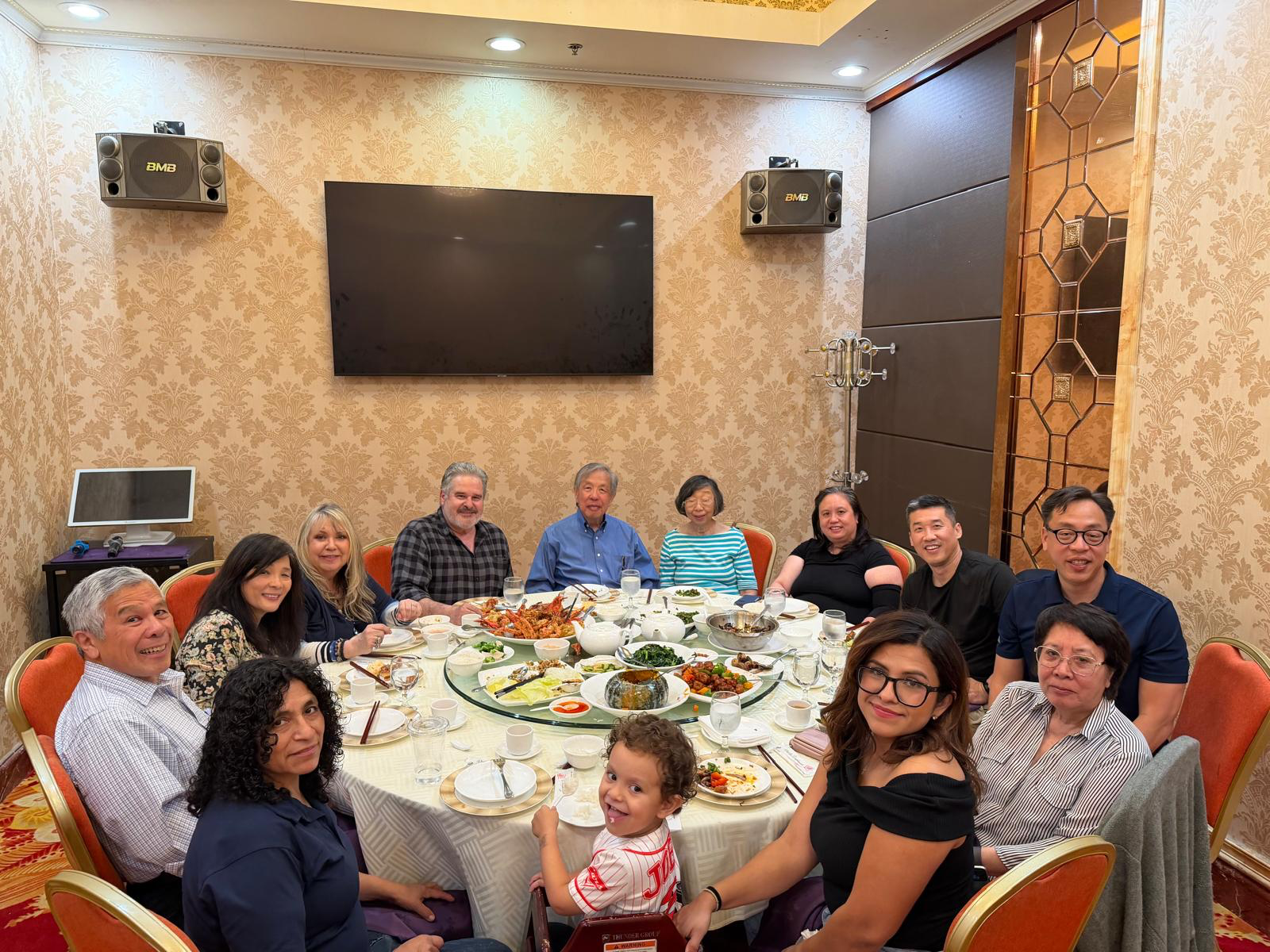 A group of diverse people gathered around a round dining table with food, in a banquet hall with a large television screen on the wall.