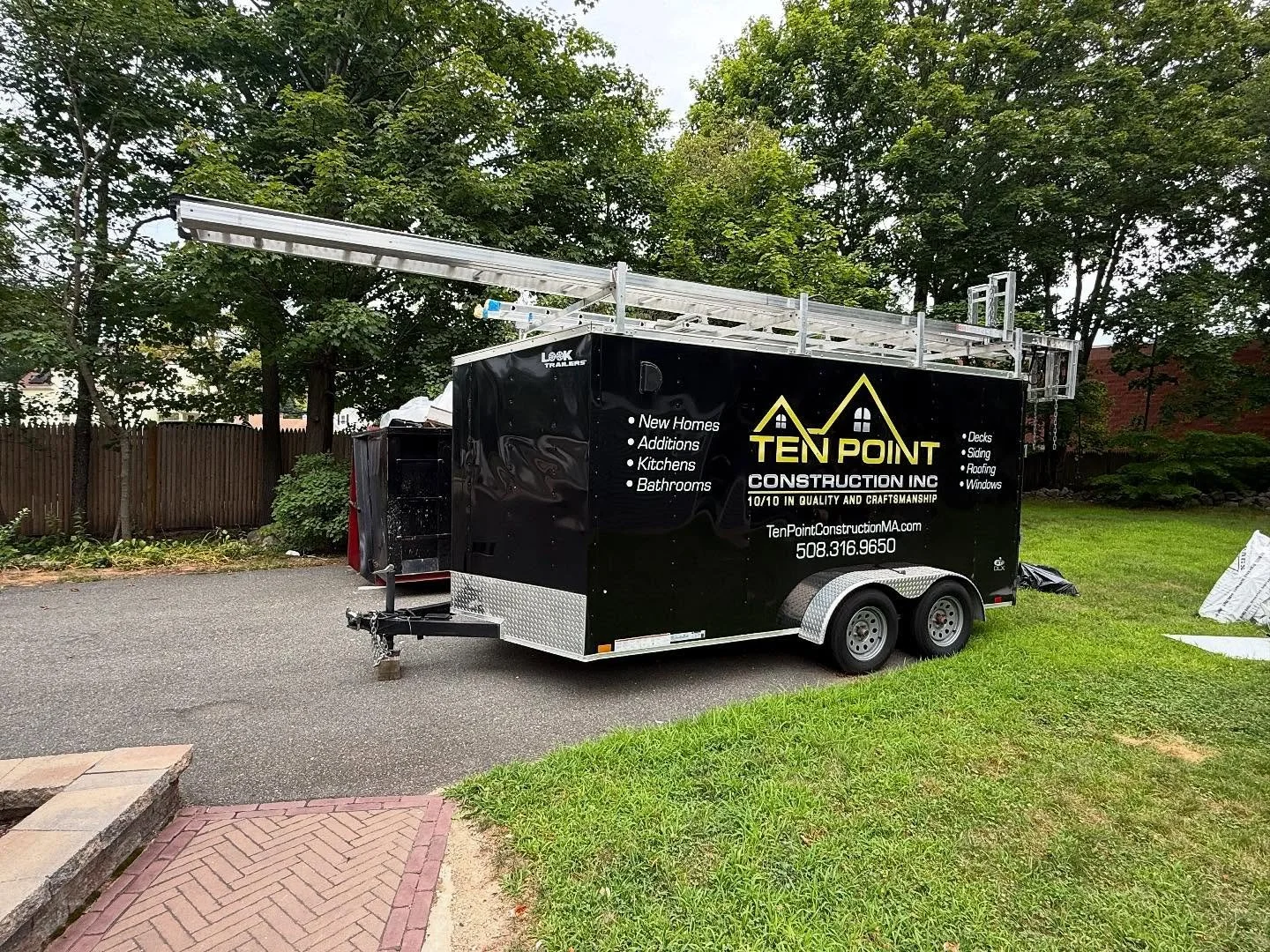 Black construction trailer parked on a driveway with a ladder on top and a wooden fence with trees in the background.