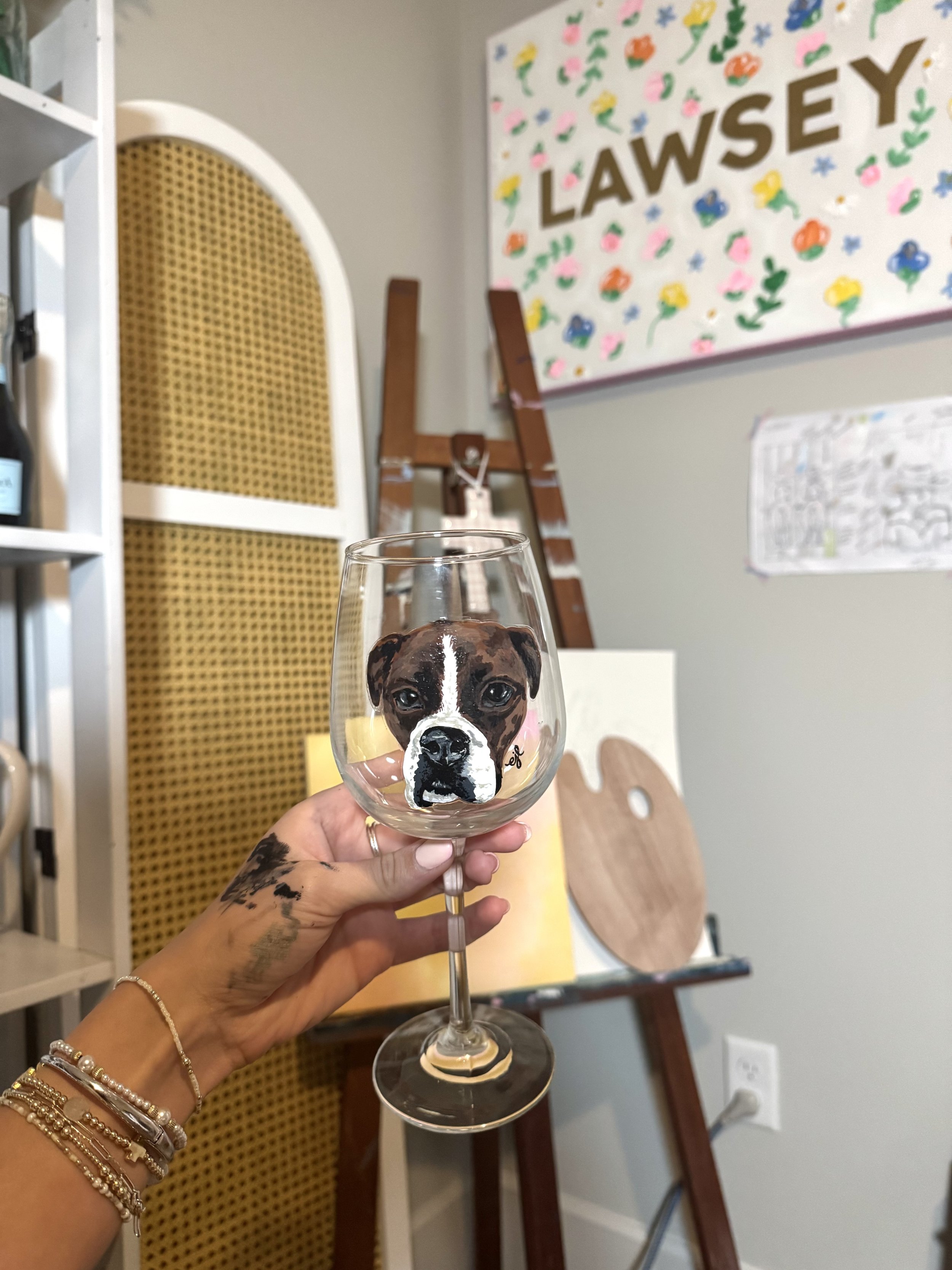 A hand with multiple bracelets holding a wine glass with a painted picture of a brown and white puppy dog face, set against a background of an art studio with a painting on the wall and art supplies.
