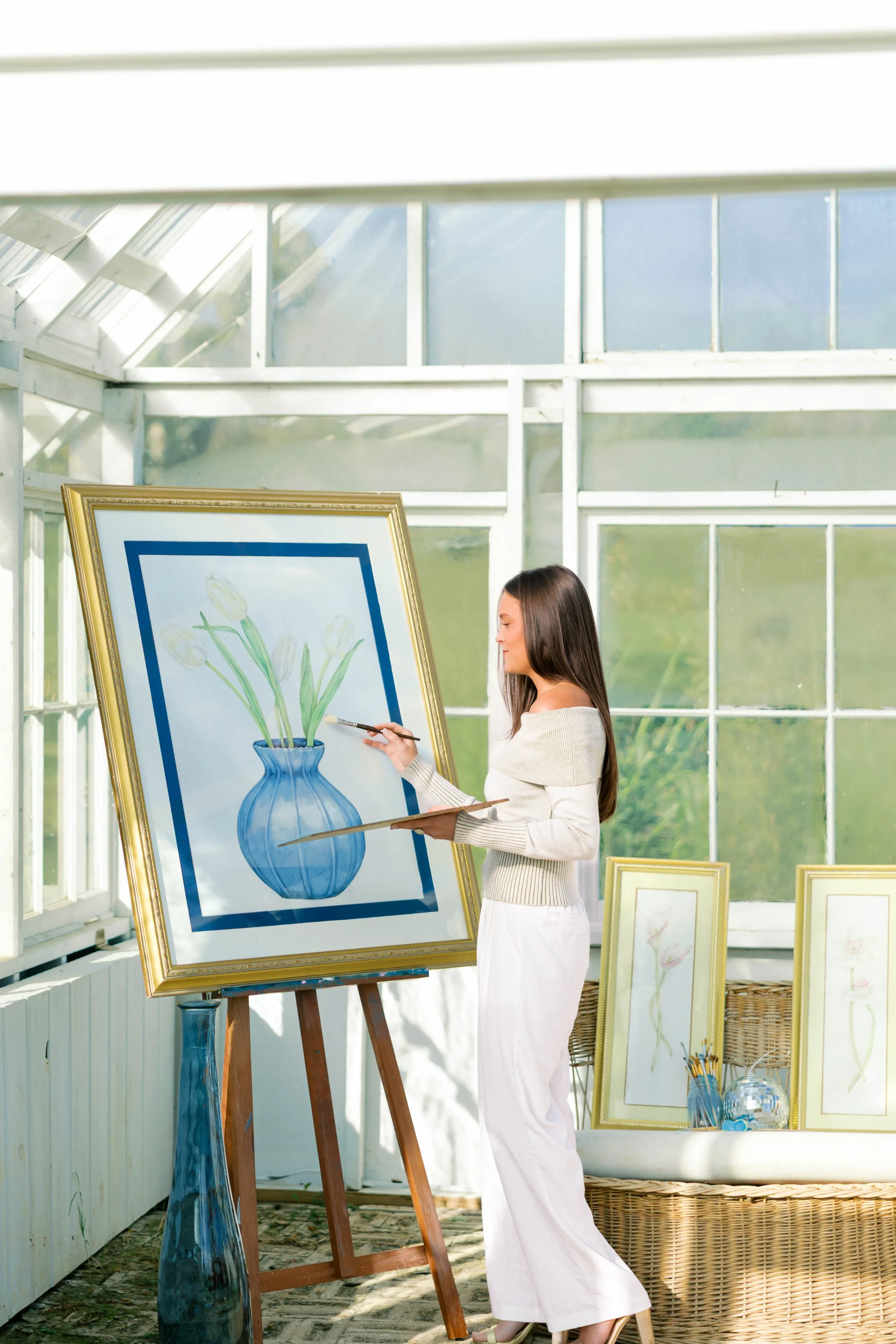 A woman painting a watercolor of white tulips in a blue vase on an easel inside a sunlit greenhouse, with framed artwork and art supplies around her.