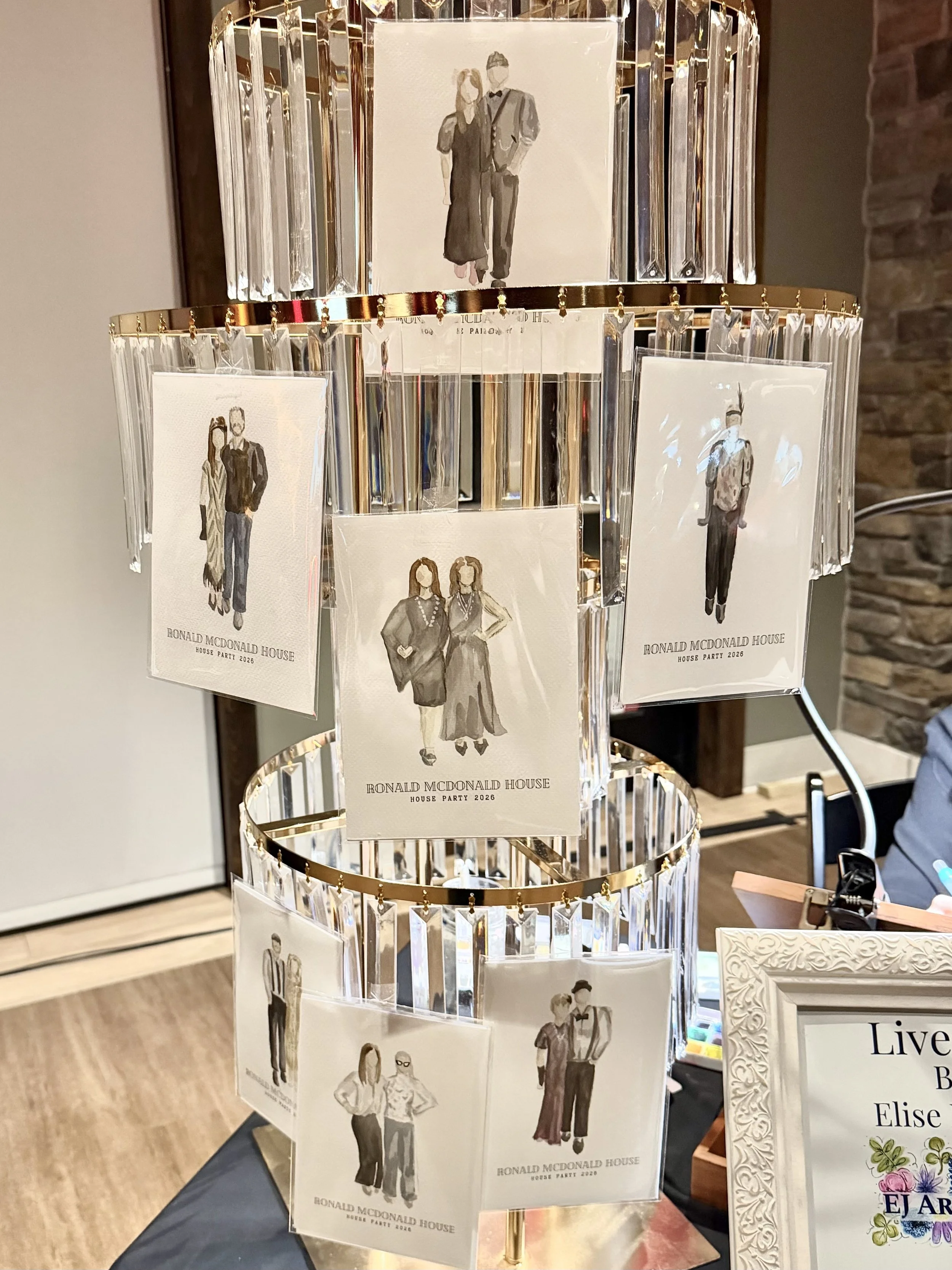 A rotating display stand with multiple cards featuring watercolor illustrations of people, labeled with "Ronald McDonald House" and "House Party 2026" at the bottom. The stand is decorated with hanging, transparent, rectangular prisms.