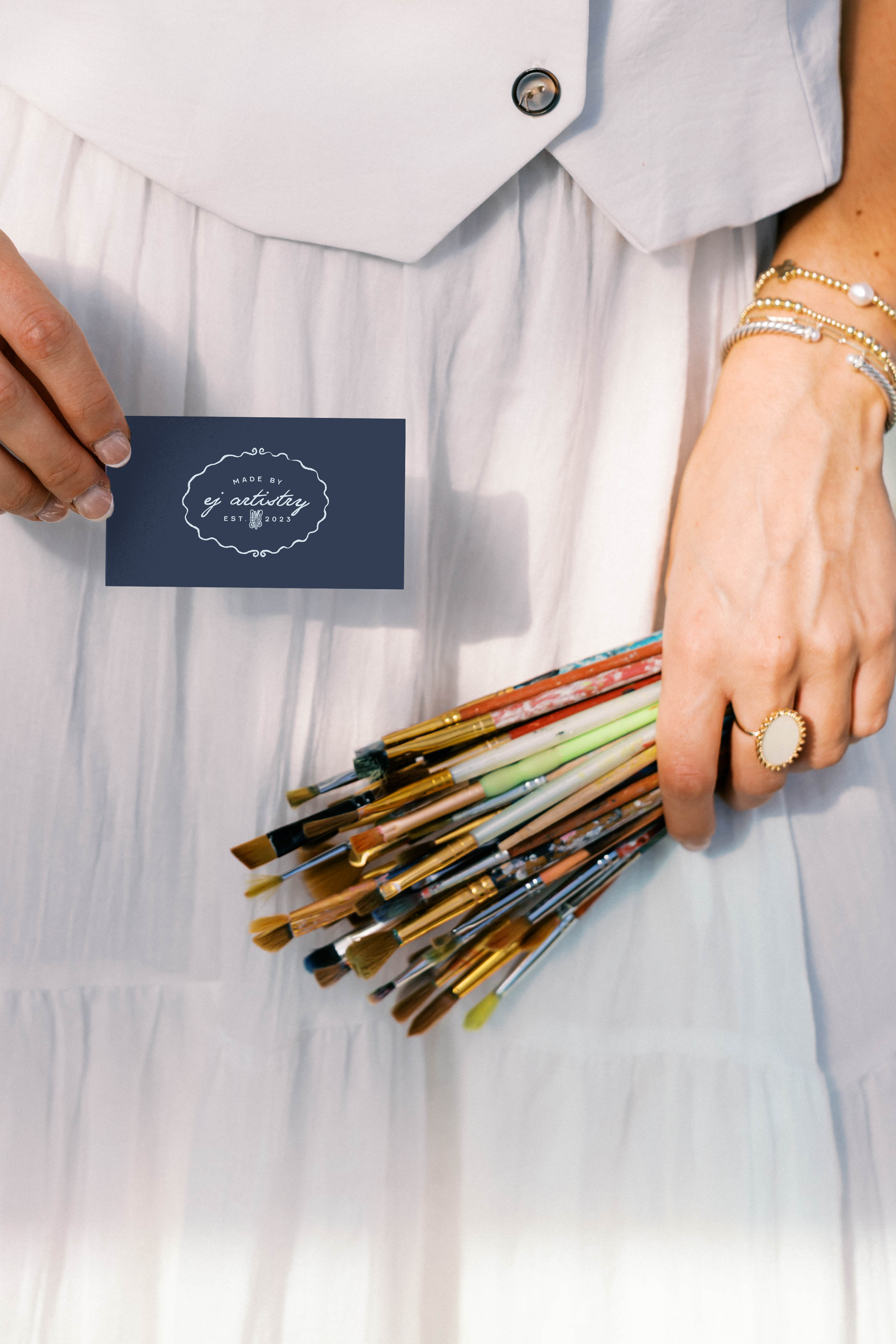Close-up of a person's hand holding a navy business card with white text and decorative border, standing next to a bundle of paintbrushes on a white fabric surface, with jewelry visible on the person's wrist and finger.