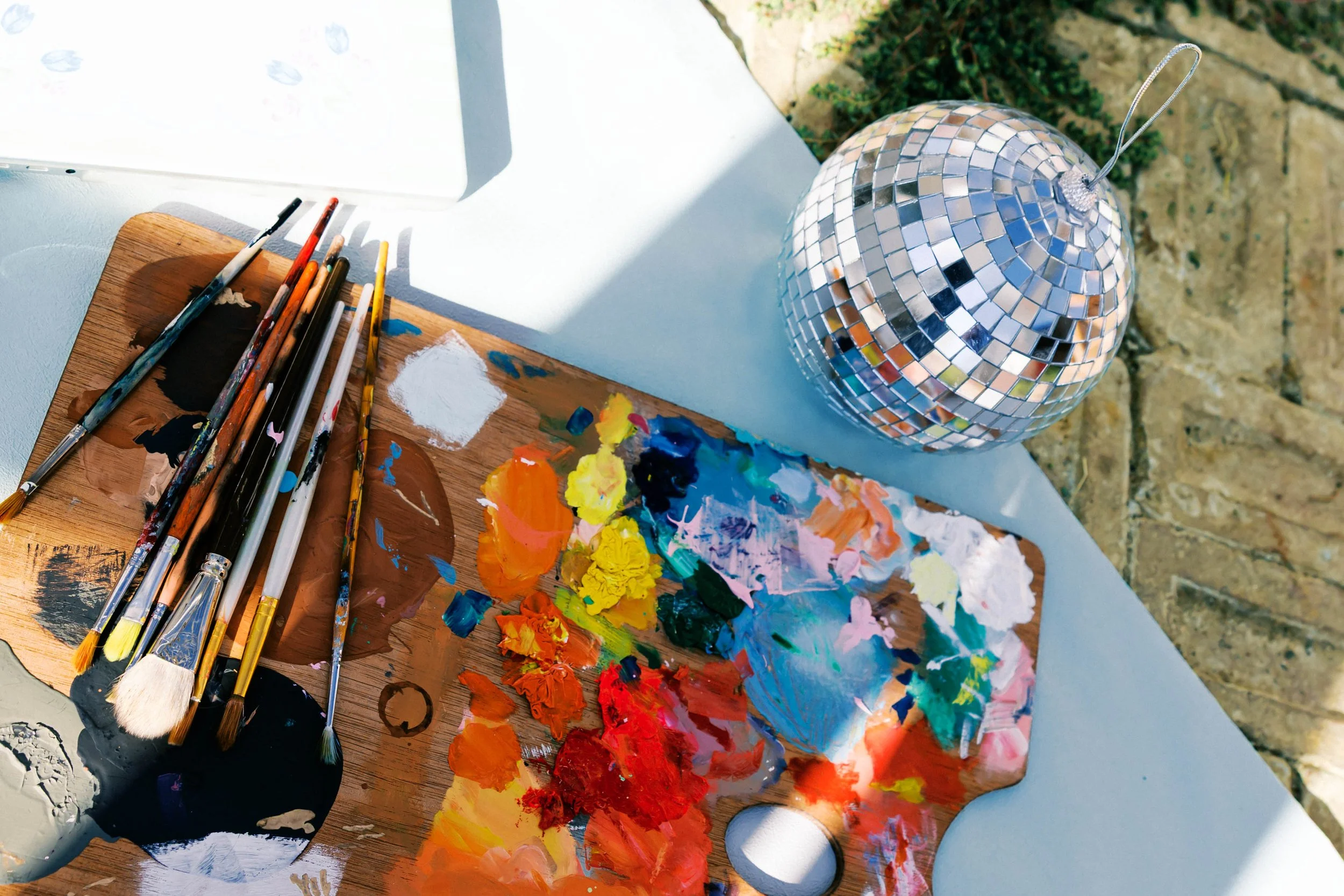 Paint palette with mixed colors, paintbrushes, and a small disco ball on an outdoor table.